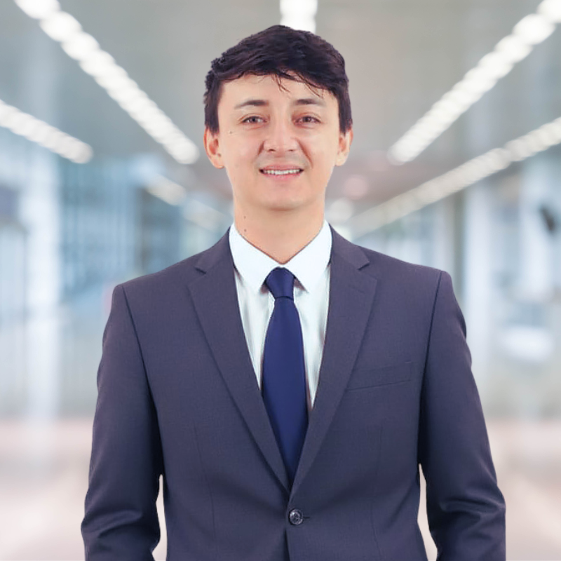 A young man in a suit standing in a modern office building with blurred bright lights in the background.