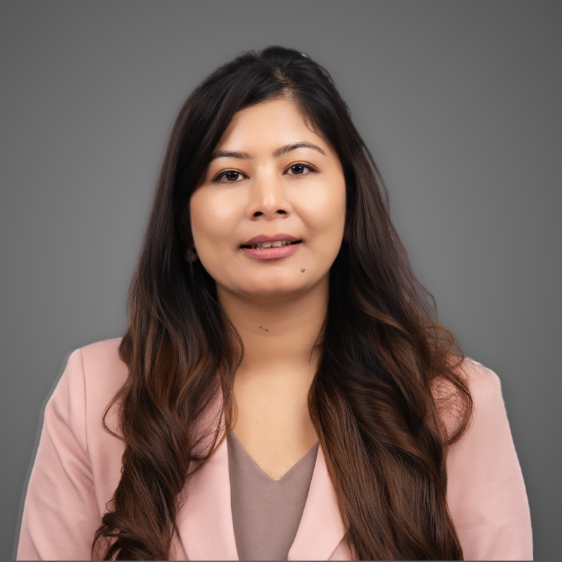 A woman with long brown hair wearing a beige blazer and white top standing in an indoor setting with glass walls and ceiling lights.