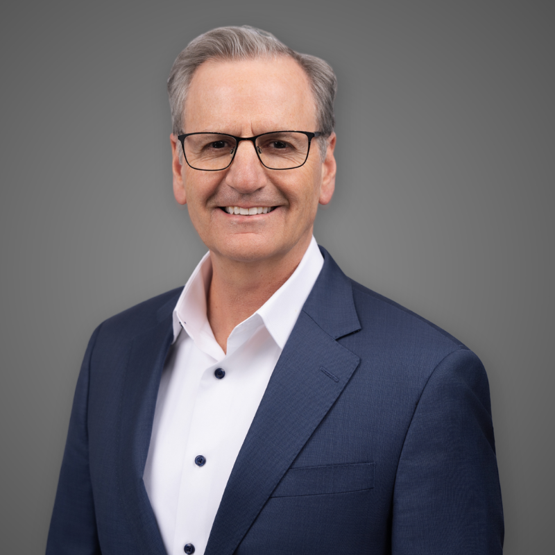 A middle-aged man with gray hair, glasses, and a friendly smile, dressed in a dark suit, white shirt, and patterned red and gray tie, standing in a modern, brightly lit indoor space with blurred background.