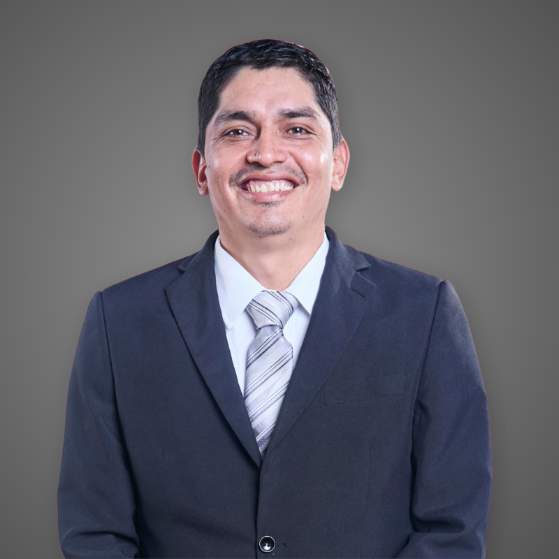 A man in a plaid shirt standing in a well-lit indoor corridor, smiling at the camera.