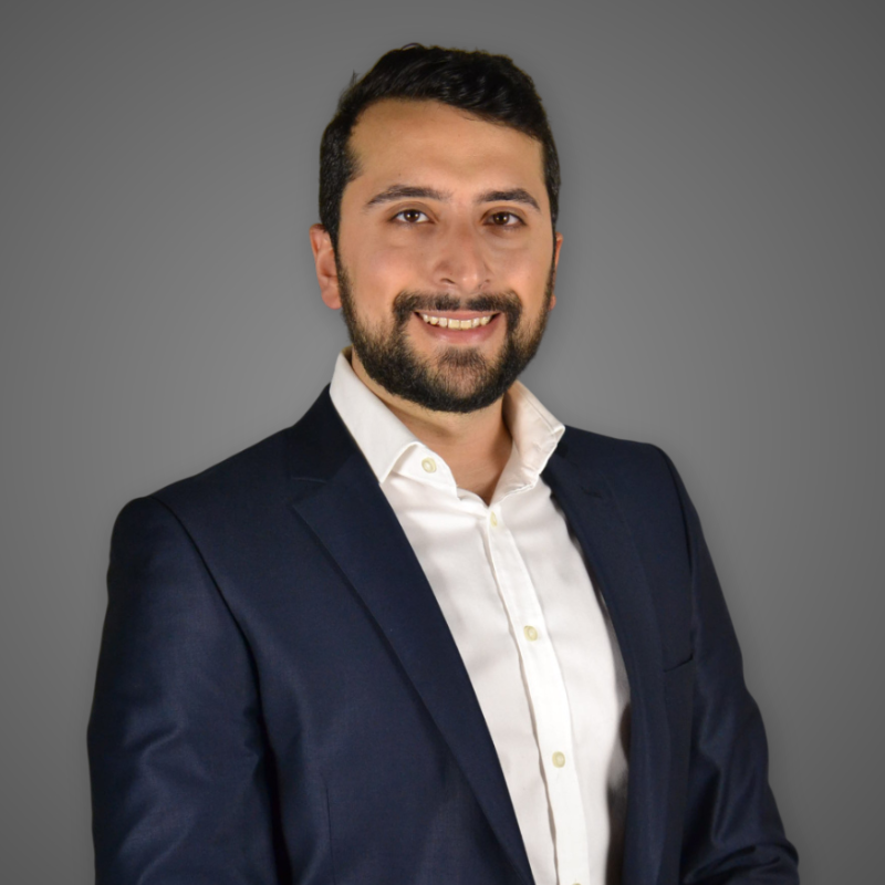 A man with dark hair and a beard, wearing a white shirt and navy blazer, smiling in a modern, well-lit indoor space.