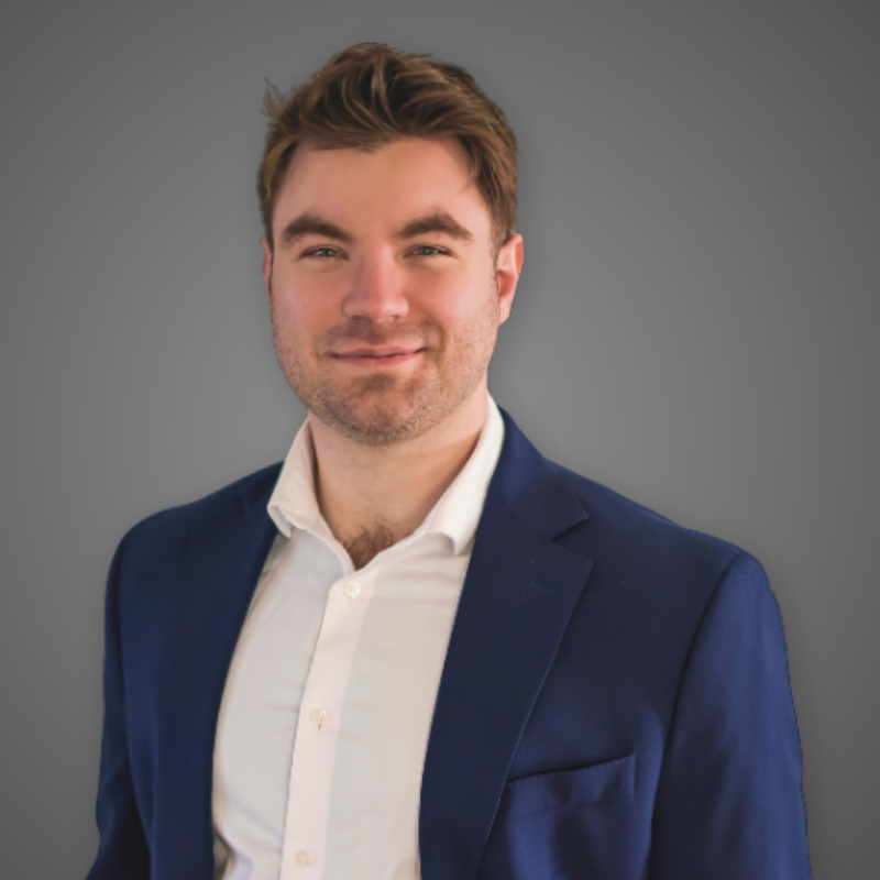 Portrait of a young man with brown hair and light stubble, dressed in a navy blazer and white shirt, smiling, standing in a modern, well-lit corridor with blurred background.