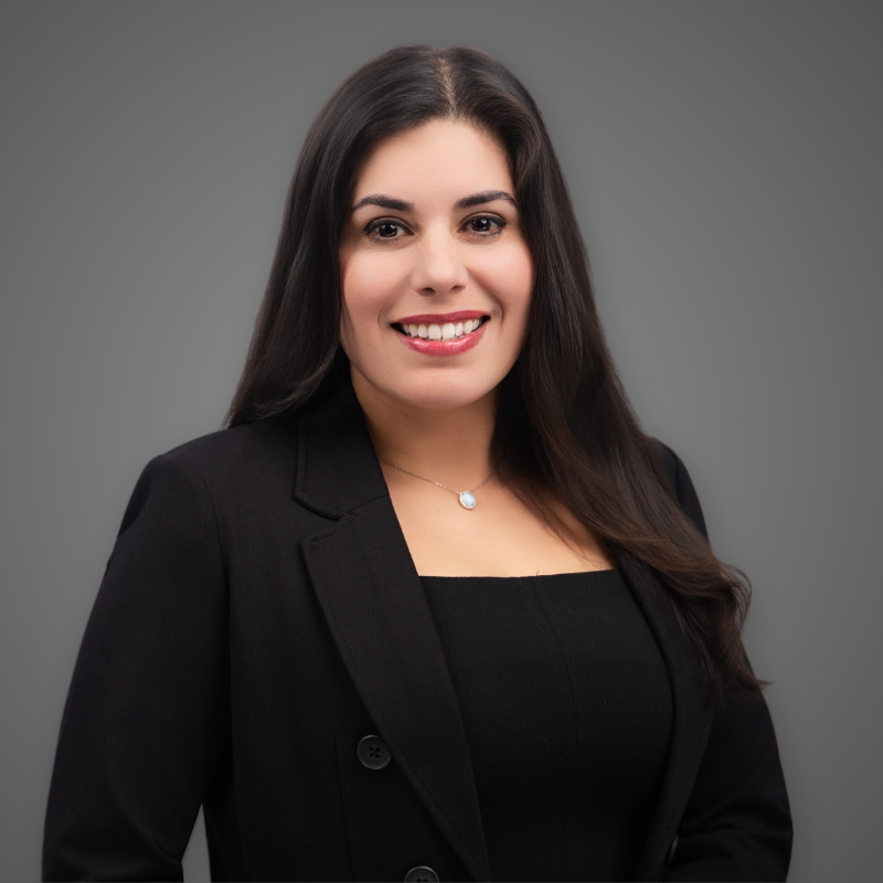 A woman with dark hair smiling at camera in an indoor setting with bright lighting and blurred background.
