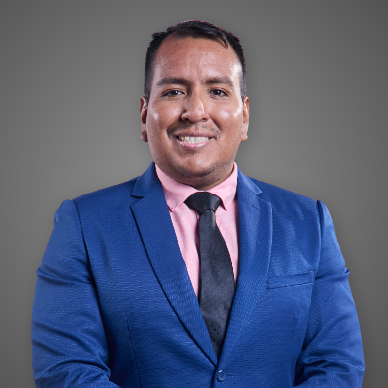 A man with glasses, short dark hair, and a goatee, wearing a dark green plaid shirt with a company logo on it, crossing his arms and smiling indoors.