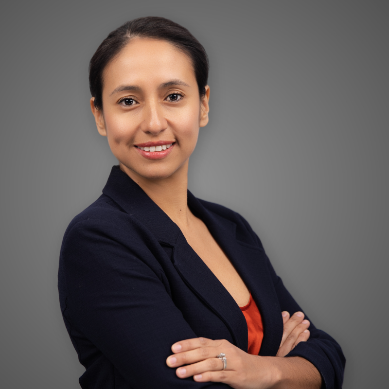 A smiling woman with crossed arms, wearing a white sleeveless dress, gold earrings, and a green necklace, standing in a brightly lit indoor space with a blurred background.