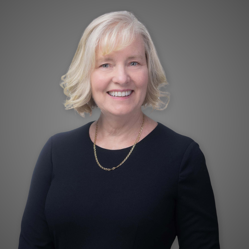 A smiling middle-aged woman with shoulder-length blonde hair, wearing a black top and a gold necklace, standing in a modern indoor setting with blurred background and bright lighting.