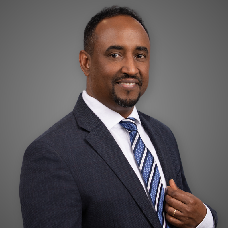 A smiling man in a suit and tie standing in a modern, well-lit corridor.