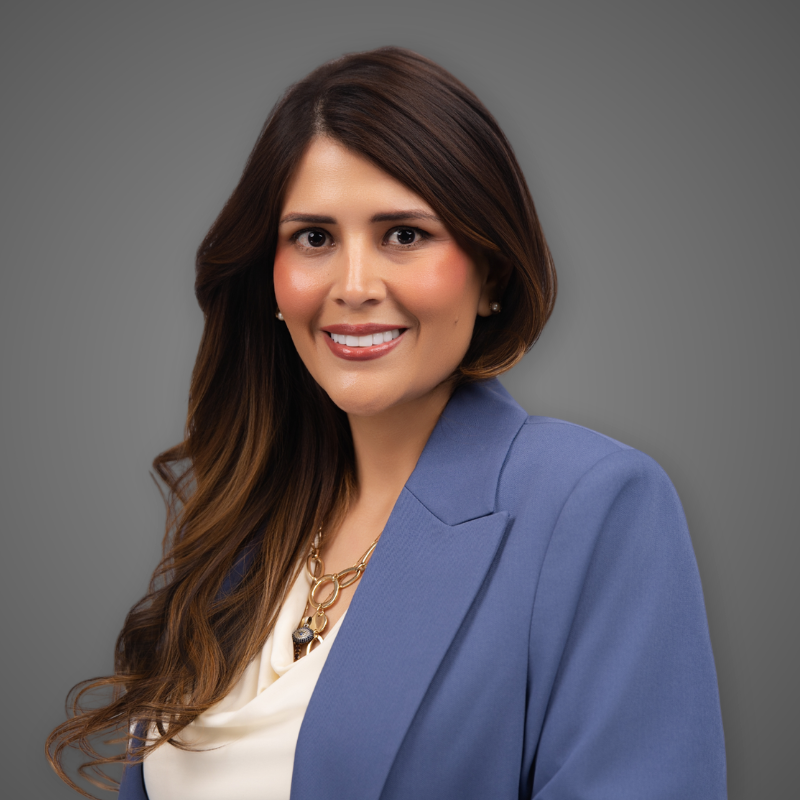 A woman with long brown hair wearing a sleeveless red dress and large earrings, smiling in an indoor setting with bright lighting.