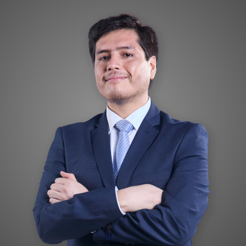 Portrait of a young man with dark hair wearing a green and blue plaid shirt, standing in a brightly lit indoor corridor with blurred background.