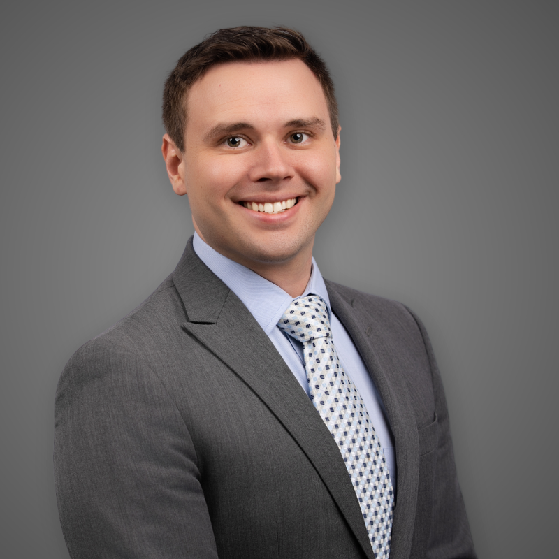 A young man in a gray suit, purple shirt, and purple plaid tie, smiling in an indoor setting with blurred modern lighting and structure in the background.