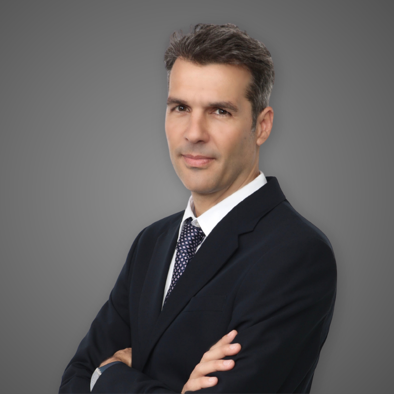 A confident man in a dark suit with a white shirt and patterned tie standing with arms crossed in a modern, bright office hallway.
