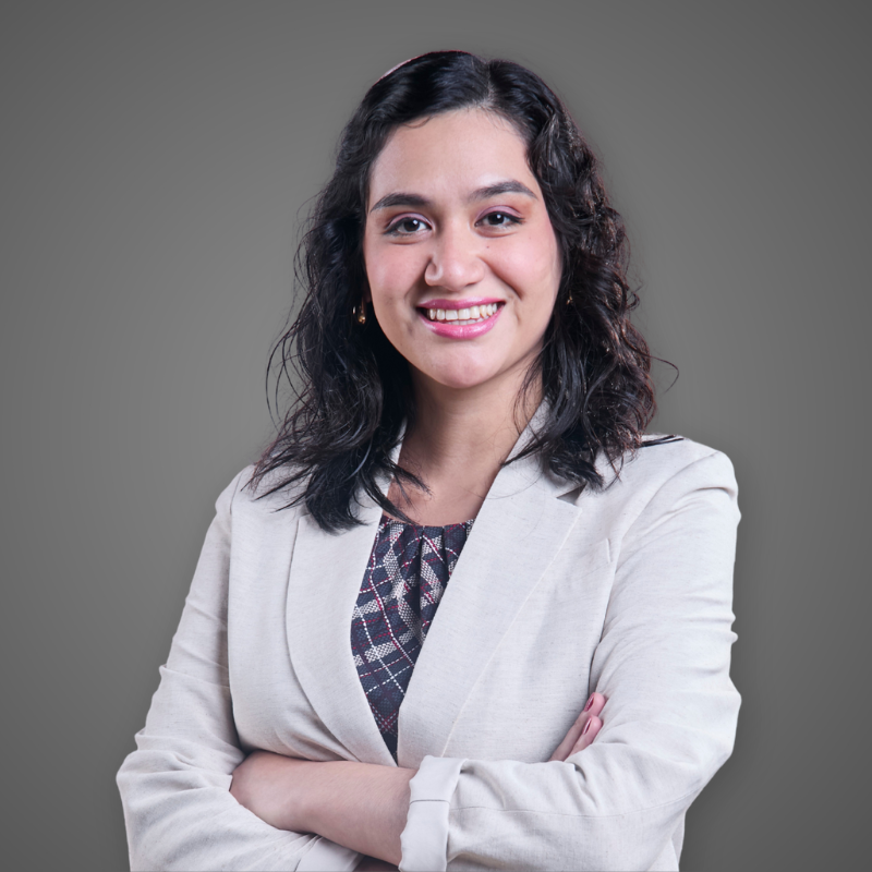 A woman with dark curly hair wearing a light blue button-up shirt with a company logo standing in a well-lit hallway.