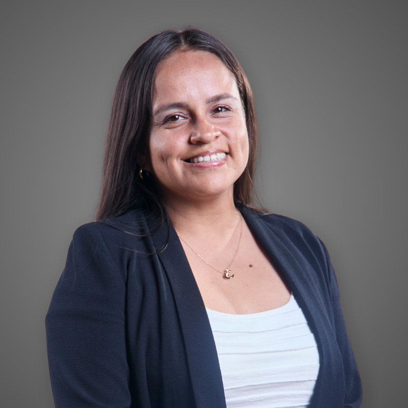 A woman with long dark hair smiling, crossing her arms, wearing a light blue collared shirt with a logo, and standing in a bright corridor.