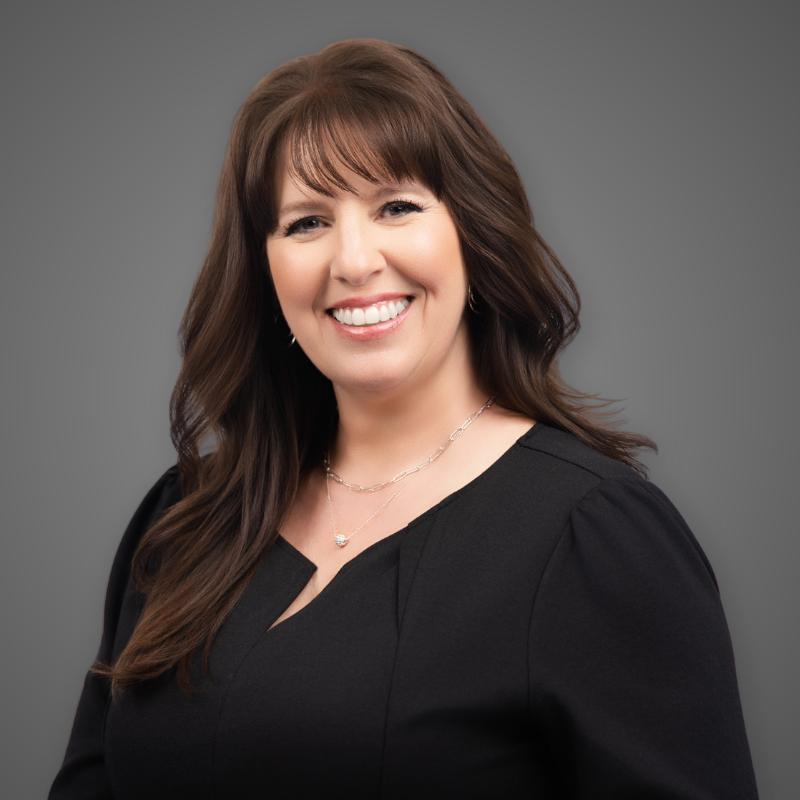 A woman with shoulder-length dark brown hair, smiling, wearing a maroon blazer over a black top, standing in a modern building corridor with bright lights and glass walls.