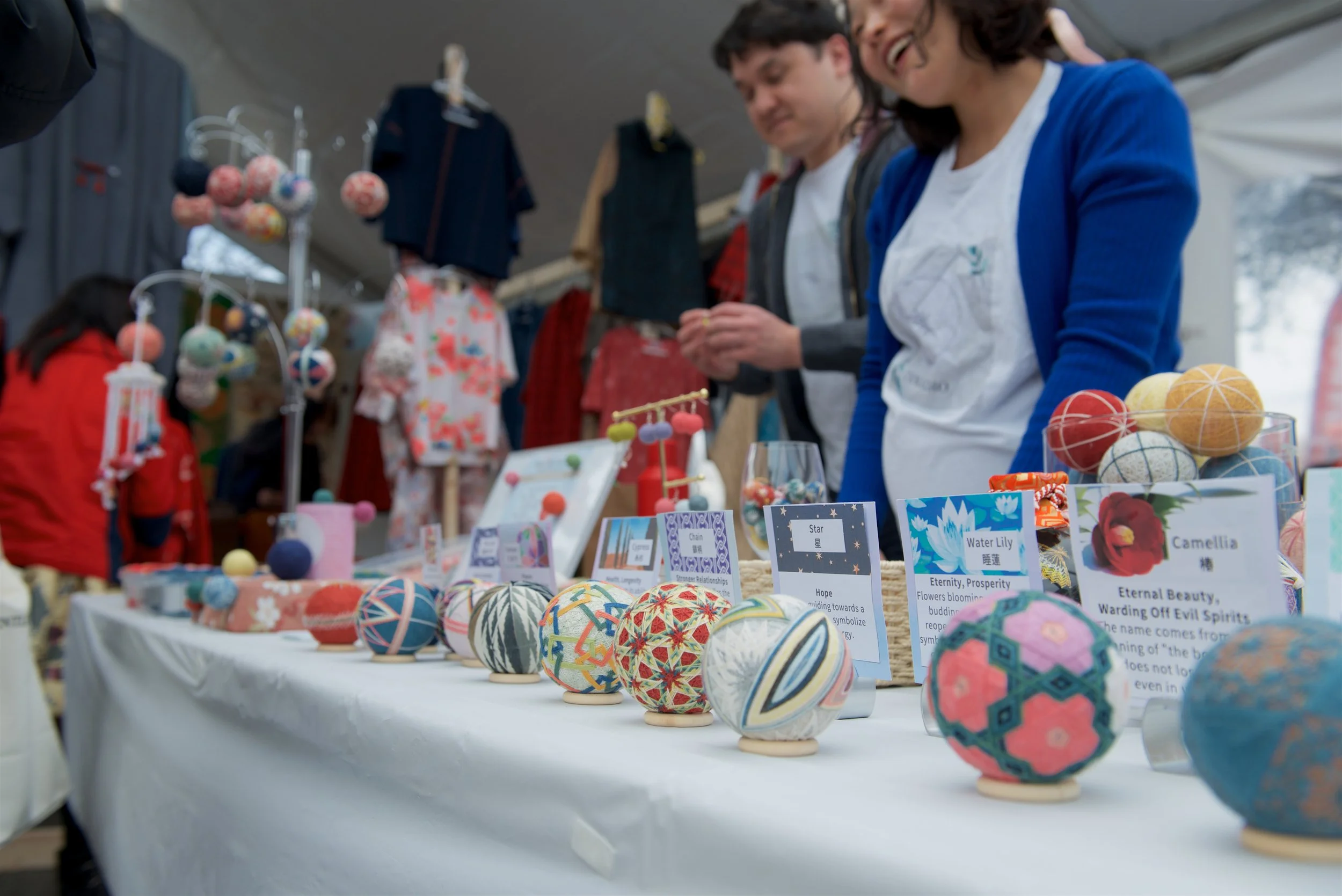 A market stall displays colorful handcrafted fabric balls and items for sale, with two people browsing and admiring the items.