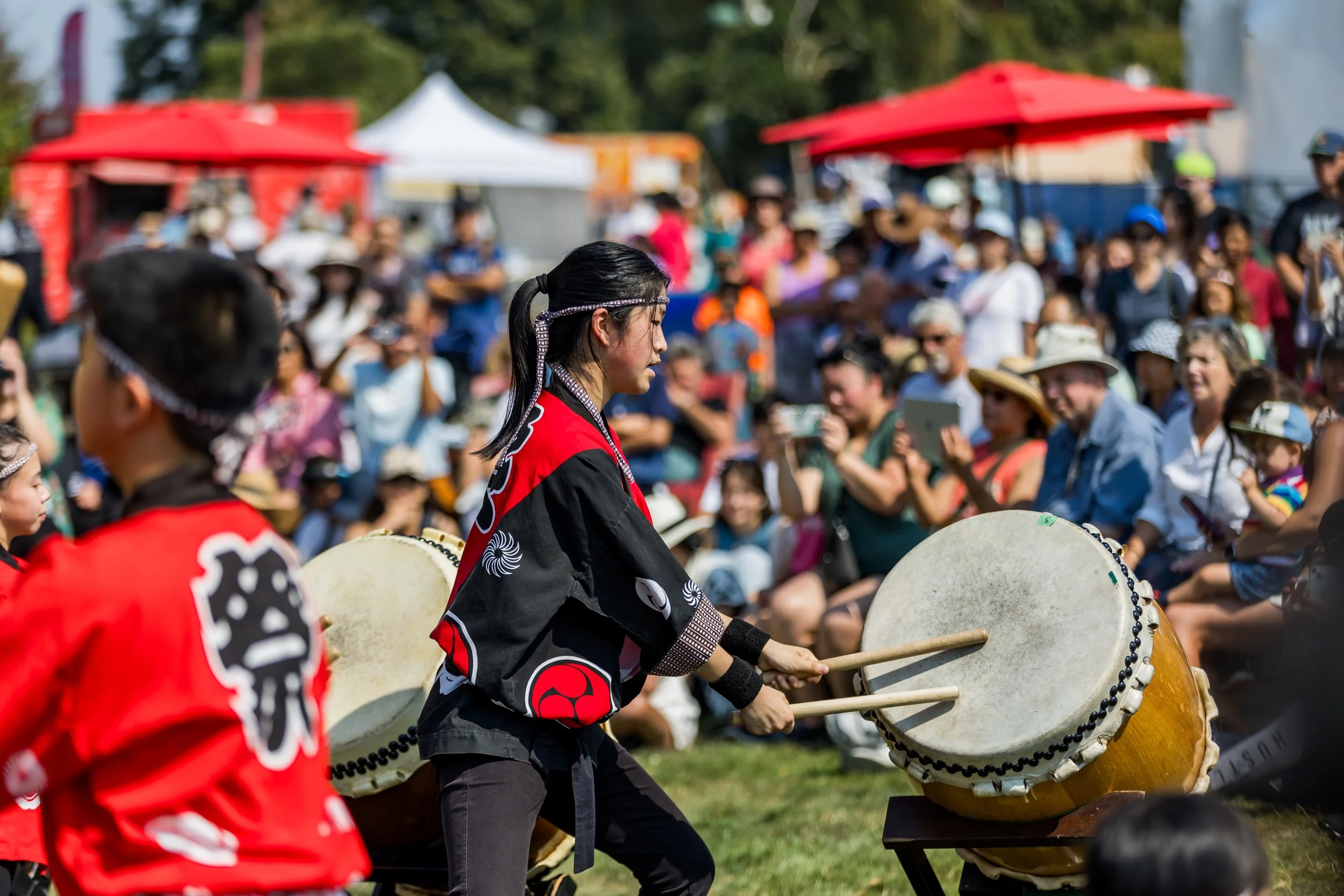 A woman playing a traditional Japanese taiko drum at an outdoor event with a crowd of spectators and festival tents in the background.