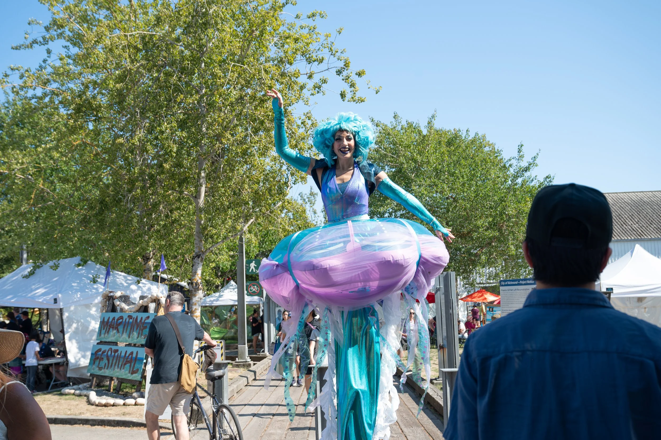 Performer dressed like a blue mermaid riding a large jellyfish sculpture at an outdoor festival, with tents and trees in the background.