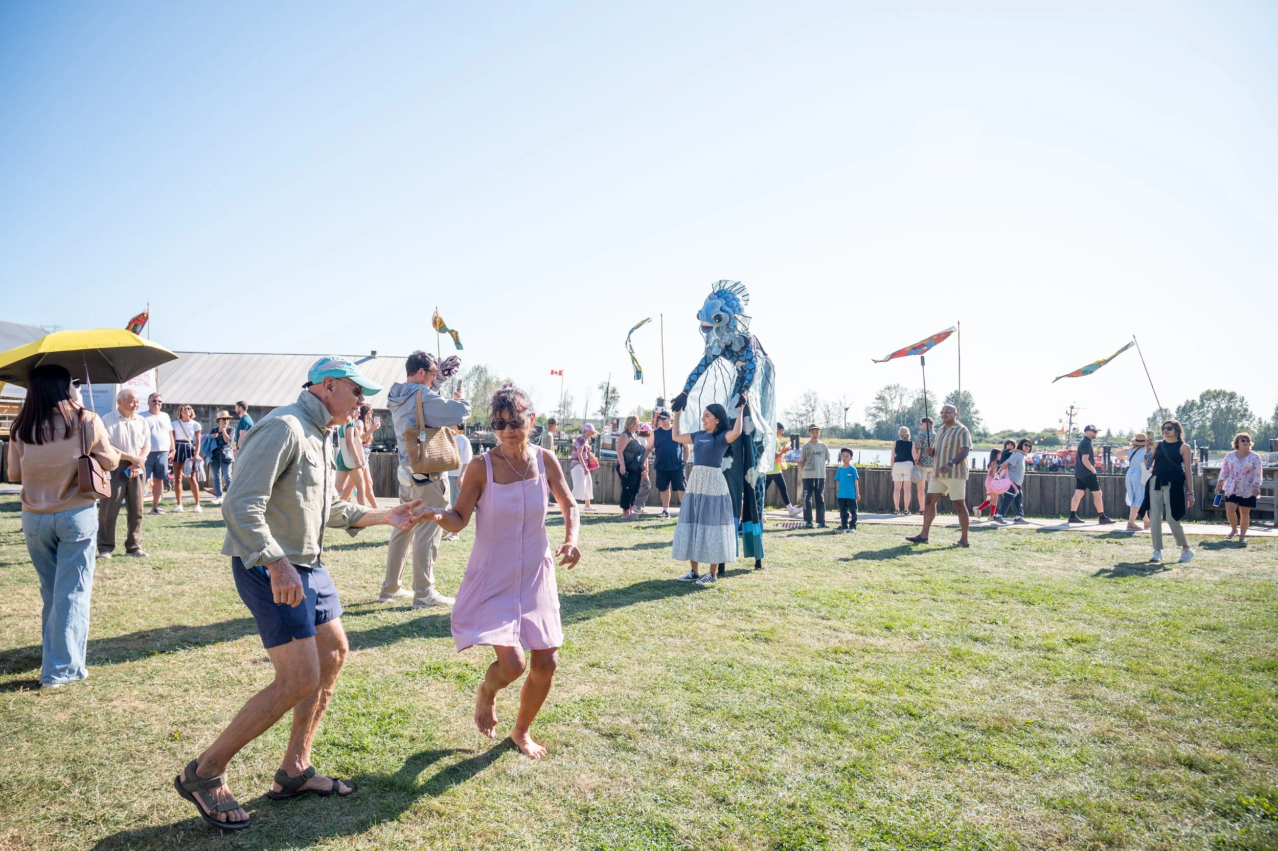 People dancing and socializing at an outdoor festival with a large blue dragon puppet and flags flying in the background on a sunny day.