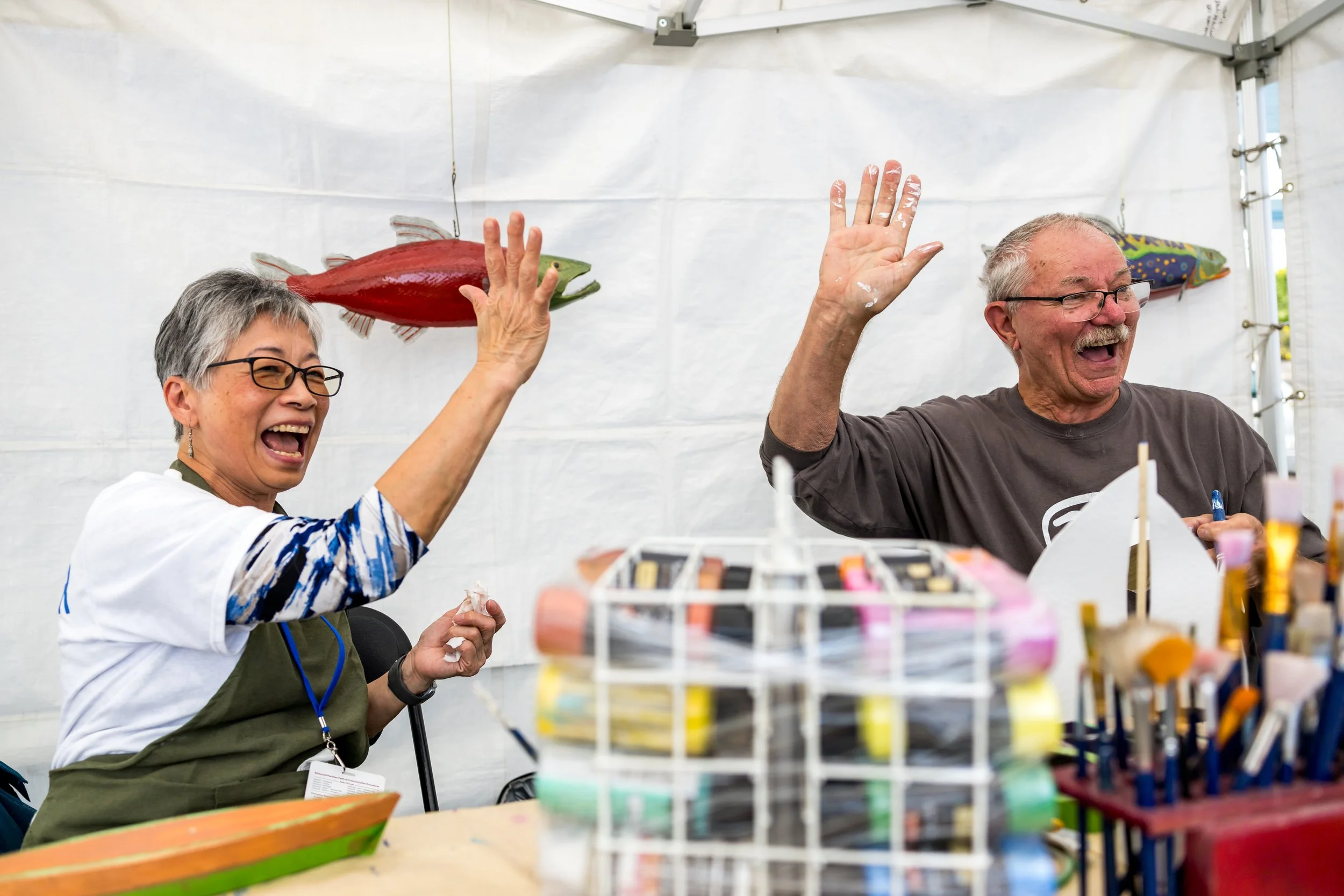 Two cheerful older adults, a woman and a man, are high-fiving at a craft booth. The woman has short gray hair, glasses, and is wearing a white shirt with blue patterns and a green apron. The man has short gray hair and is wearing a gray t-shirt with glasses. They are inside a white tent with booths displaying crafts, brushes, and paints.