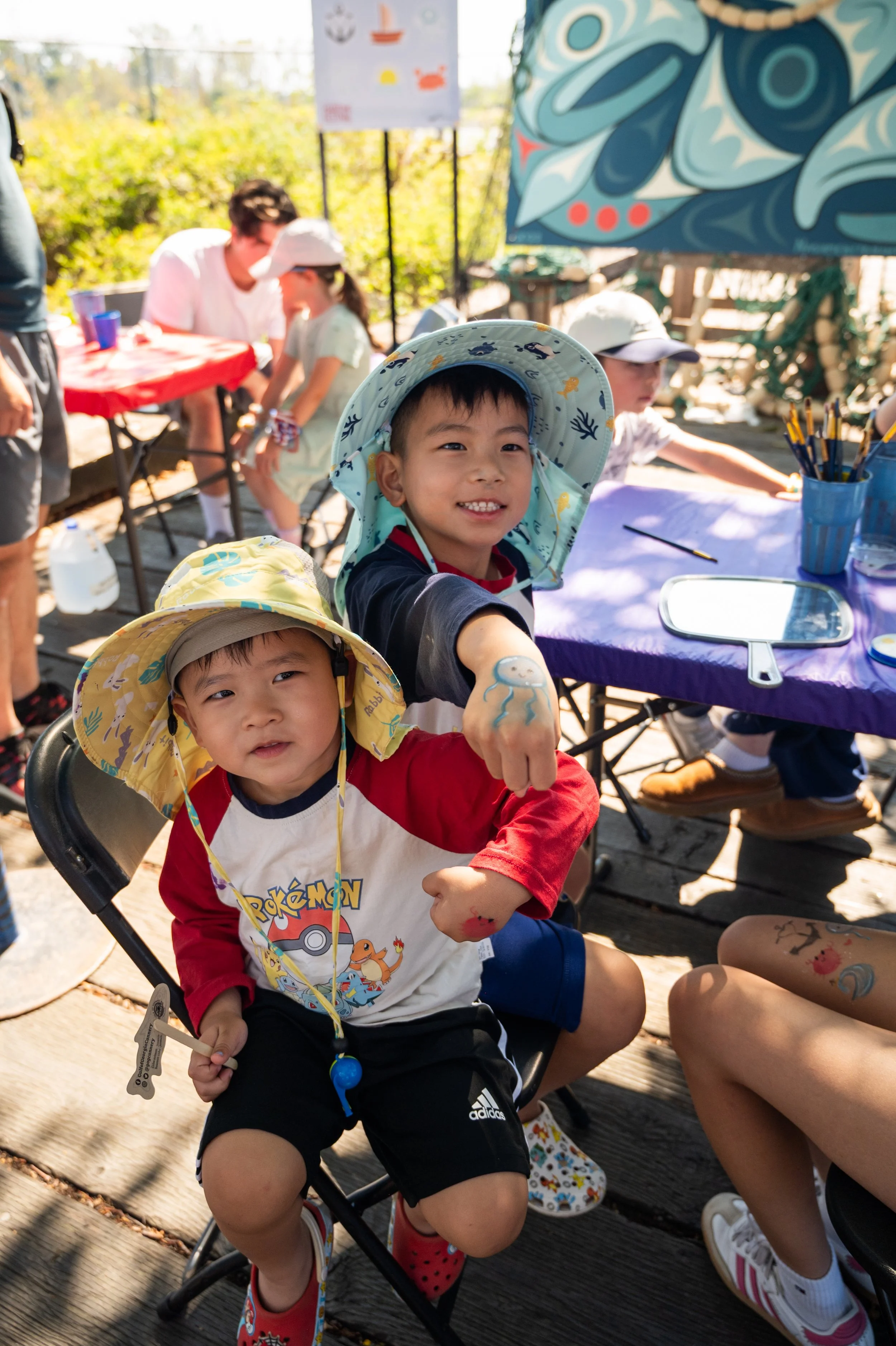 Two young boys at an outdoor event wearing hats, with one showing a jellyfish drawing on his arm and the other with the Pokémon logo shirt, seated in a chair. There are tables with supplies and people in the background.