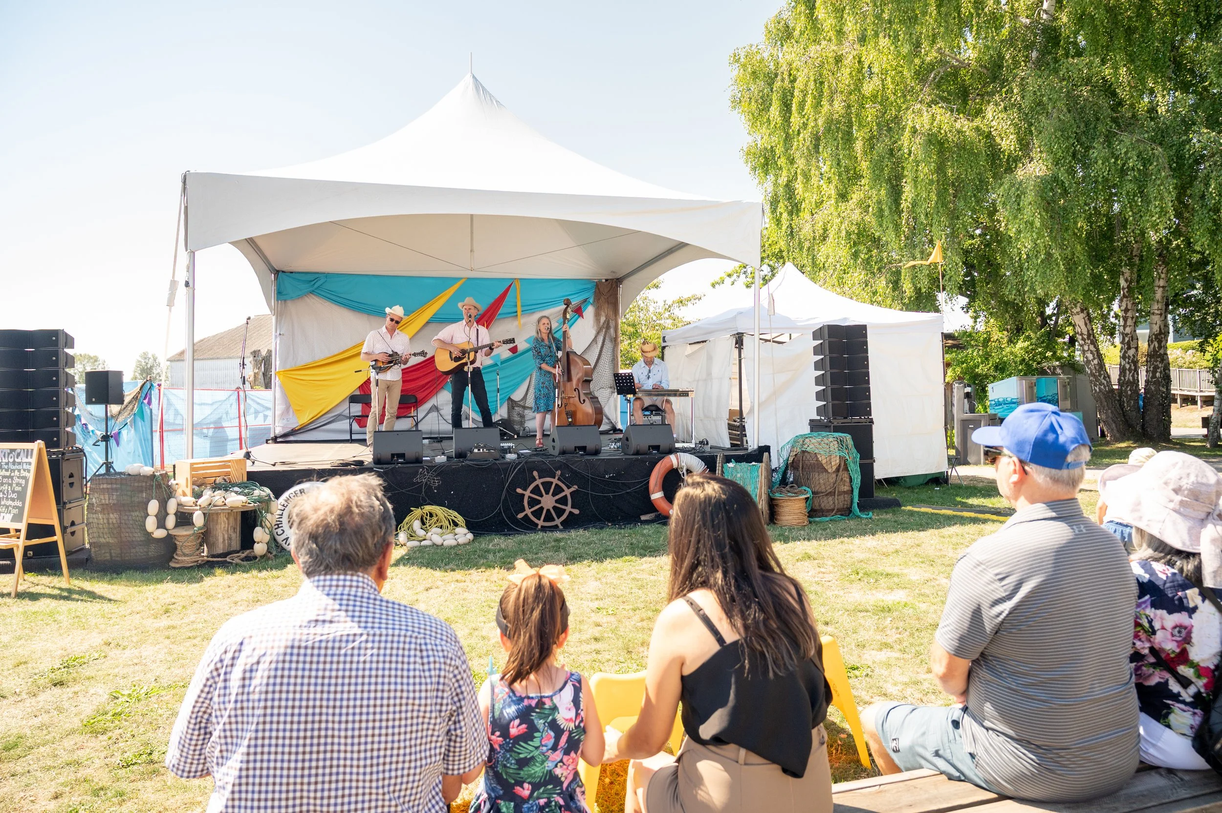 A live outdoor band performing on a stage with four members, including a woman singer, in a park on a sunny day, with an audience sitting on the grass and benches.