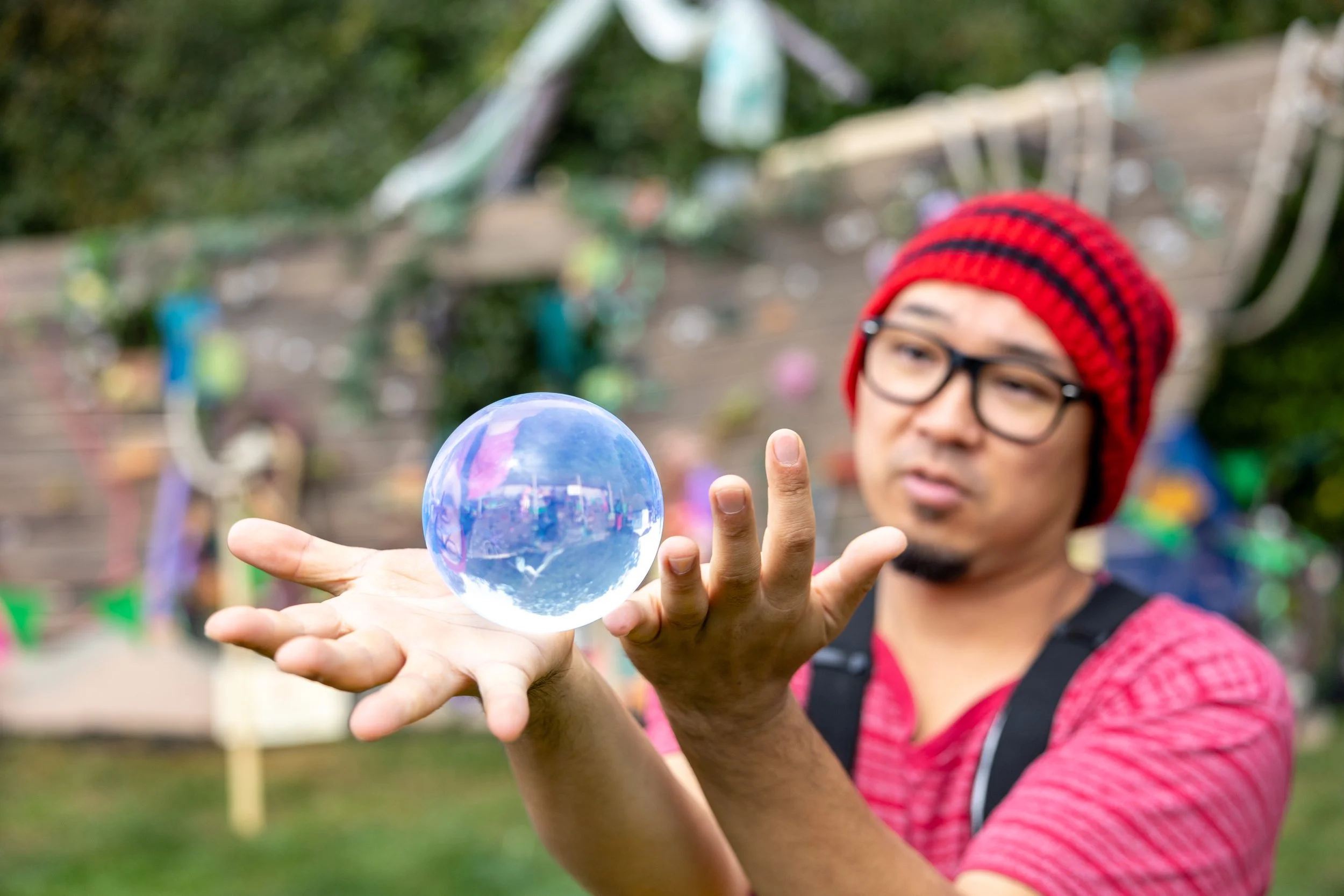 A man with glasses, a red and black beanie, and a red shirt holds a glass sphere in his hand outdoors with a wooden fence and colorful decorations in the background.