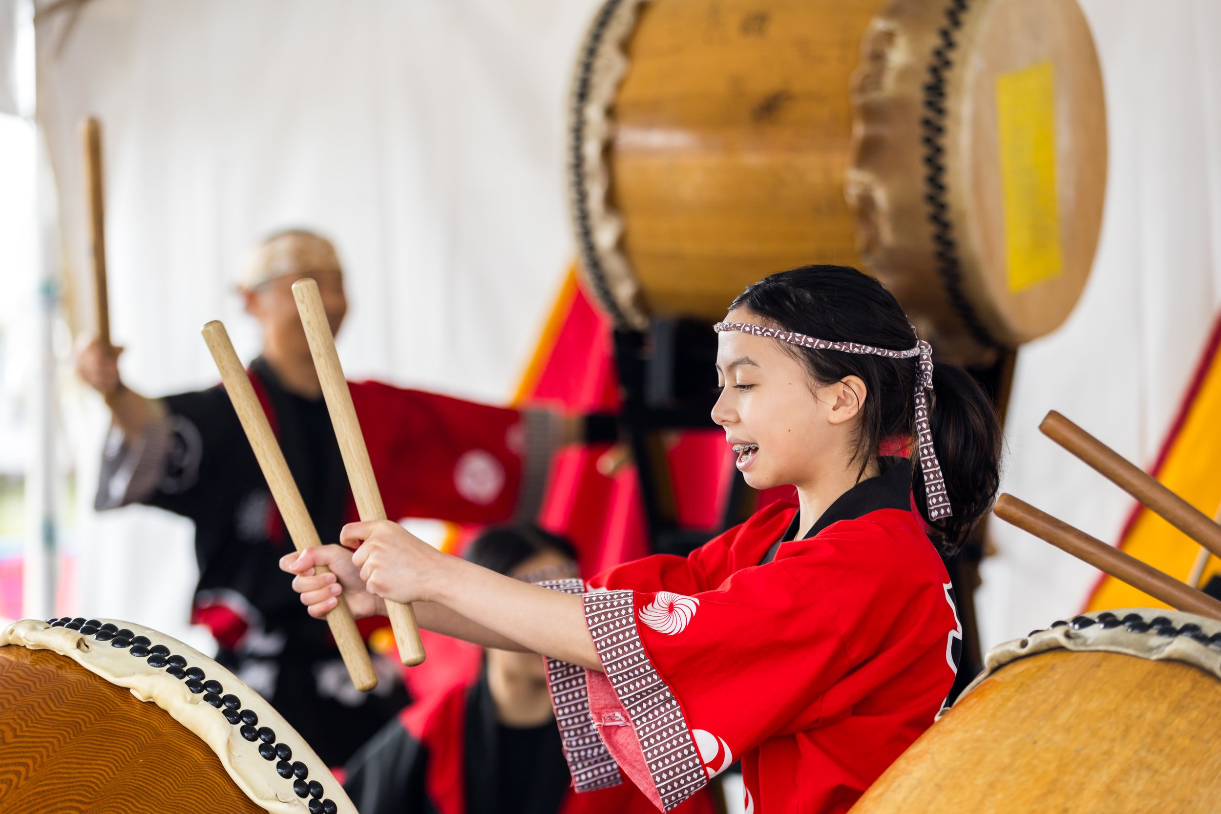 Young girl playing a large traditional drum, wearing a red and black outfit with a patterned headband, during a cultural festival.
