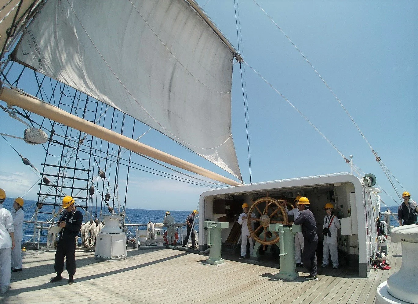 People wearing yellow hard hats operating and standing on a ship's deck with sails, ropes, and a large wheel, under a clear blue sky.