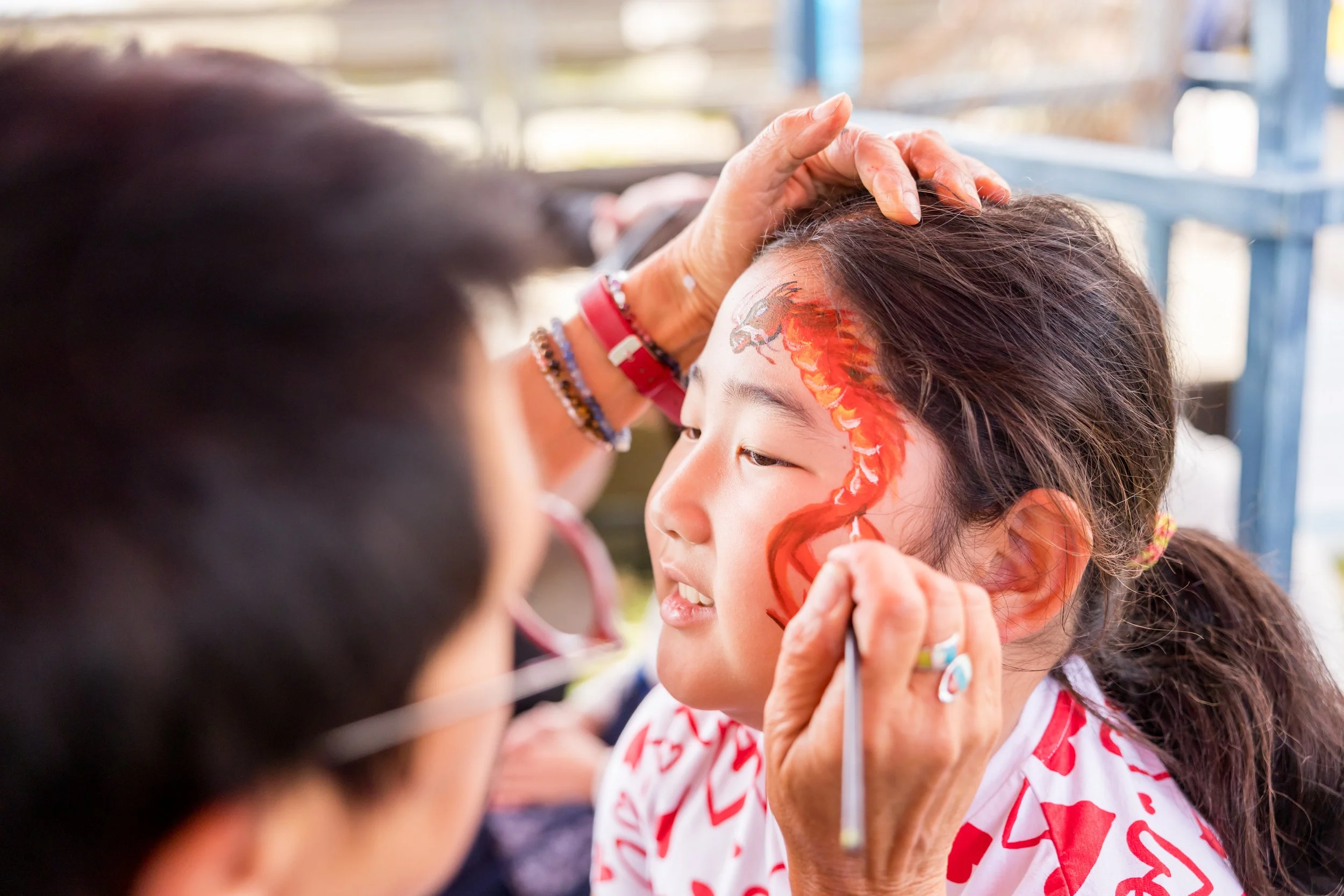 A young girl with a painted dragon on her face being painted by an artist.