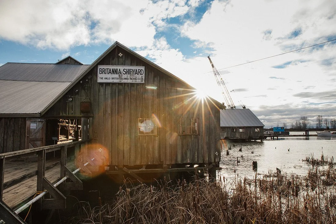 A wooden building labeled 'Britannia Shipyard' situated by the water with a dock and boat in the background, sunlight shining through the clouds.