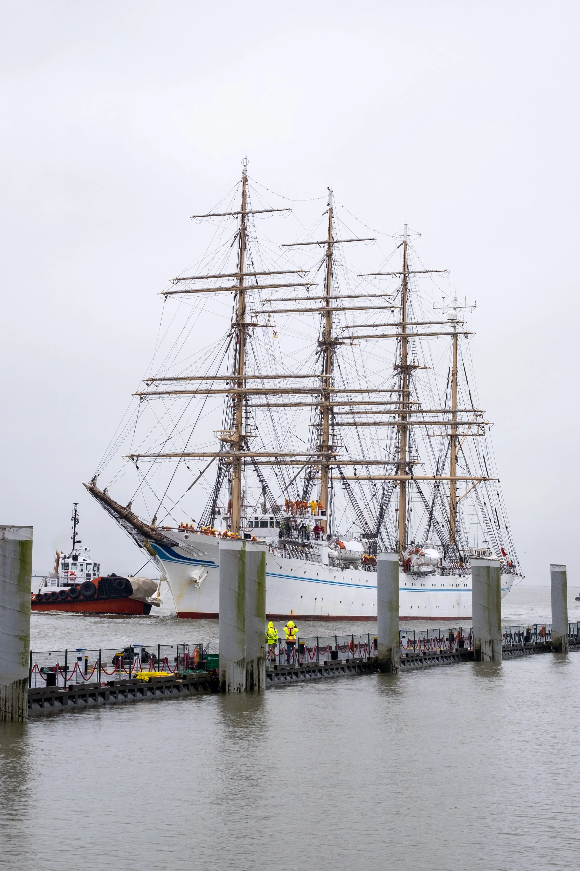 A large white sailing ship with multiple masts being guided into a harbor by a small tugboat, with workers in yellow safety vests on the dock.