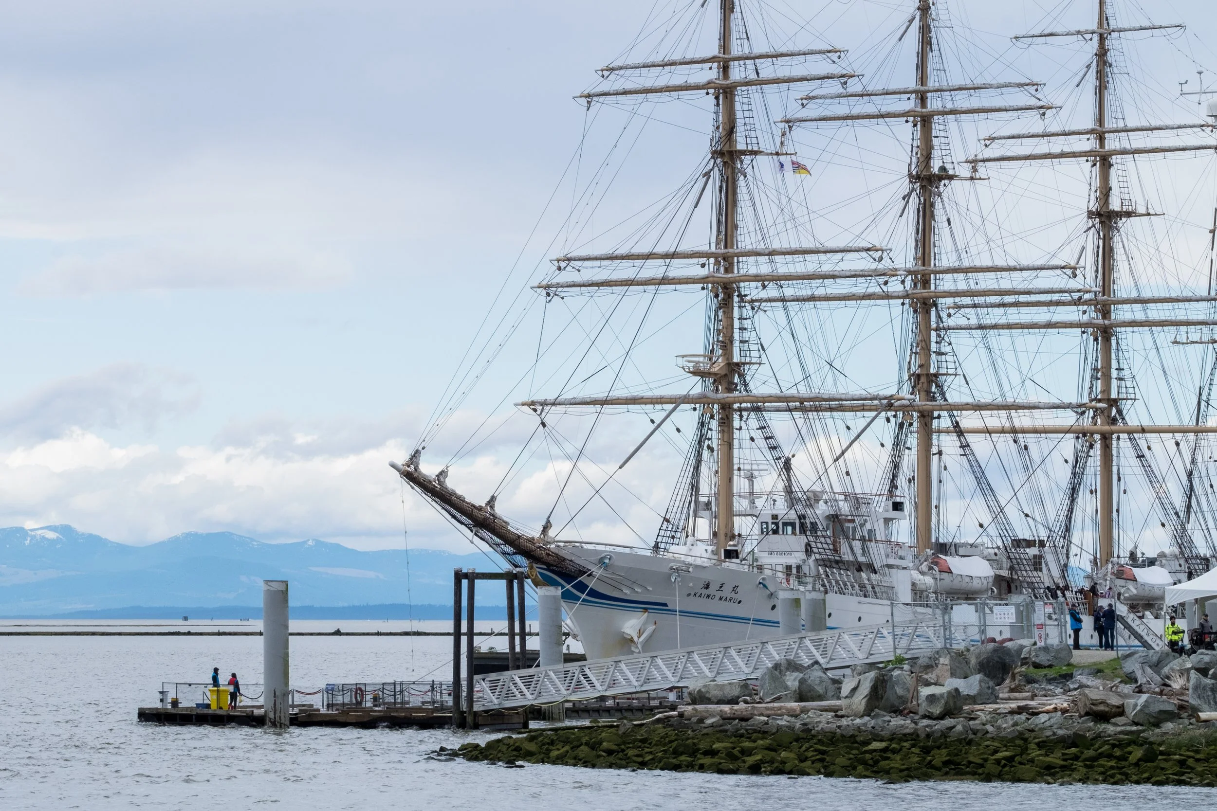 A large wooden sailing ship docked at a pier with people walking nearby, mountains in the background, and a cloudy sky.