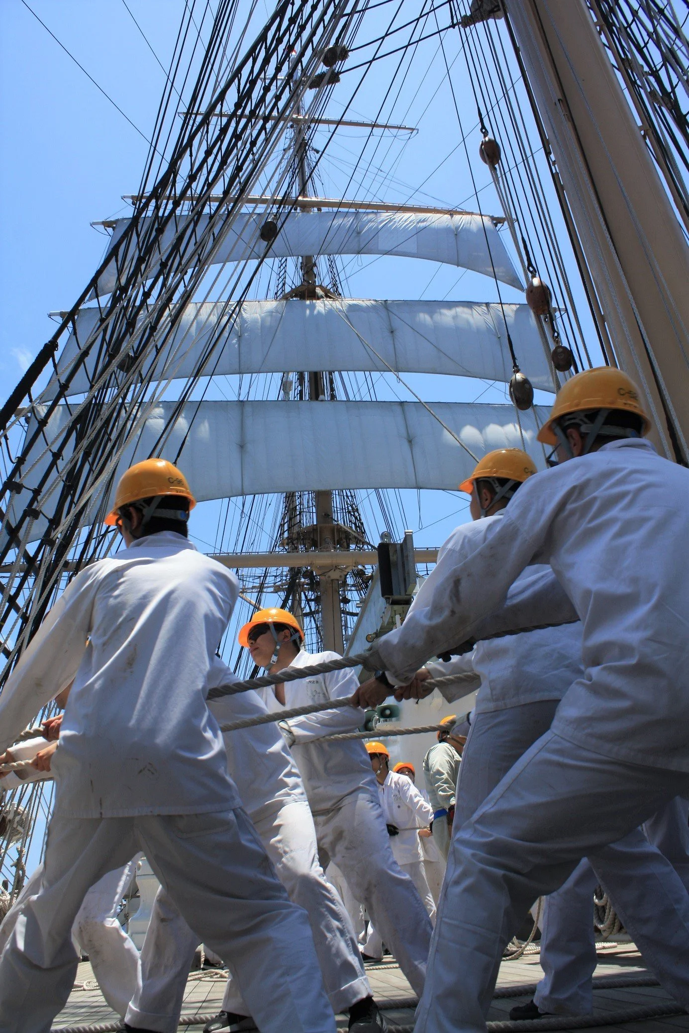 Crew members on the deck of a tall ship working together, dressed in white uniforms and wearing yellow safety helmets, with the ship's rigging and sails towering above them against a blue sky.