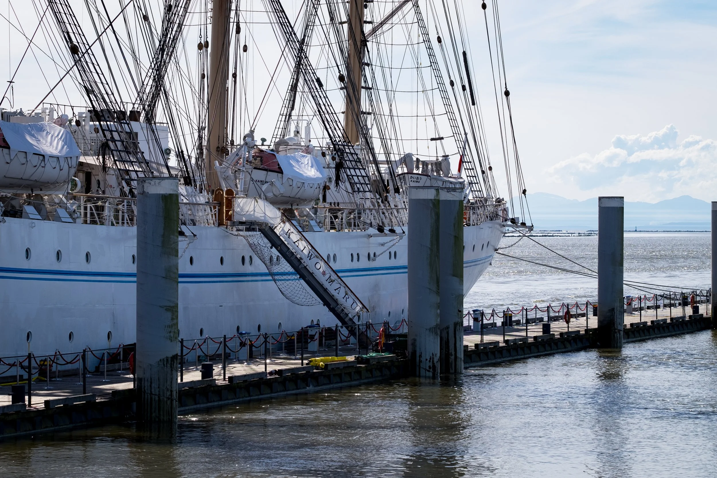Large white sailboat docked at a pier with multiple masts and rigging, alongside calm water and distant land and sky.