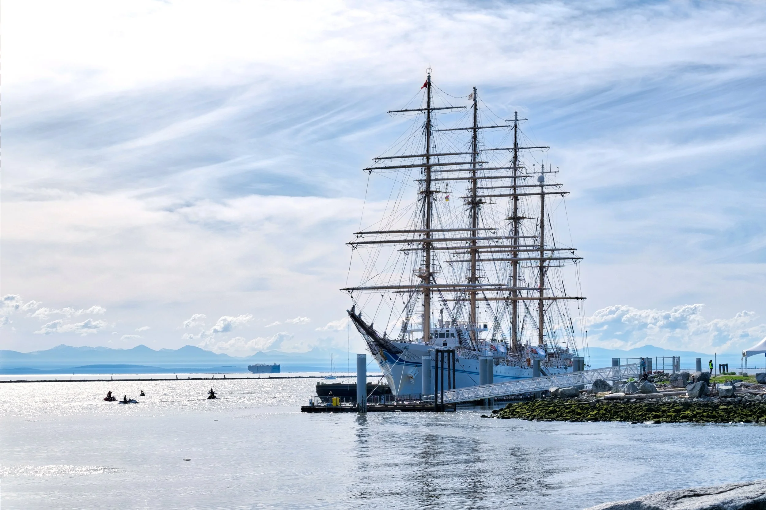 A large tall ship with multiple masts docked at a harbor, with a partly cloudy sky in the background.