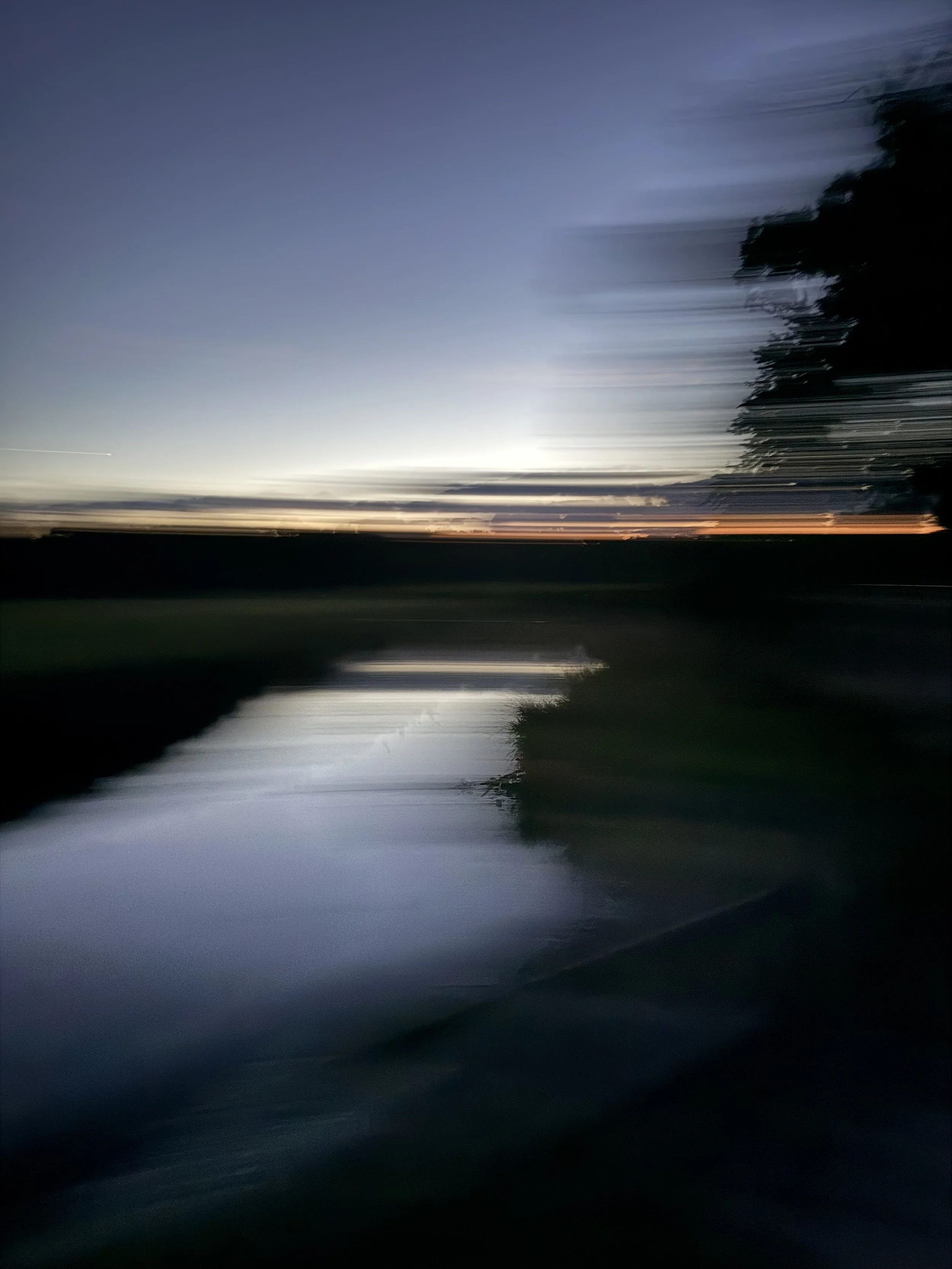 A blurred view of a creek and dramatic clouds over forest and grasslands at nightfall.