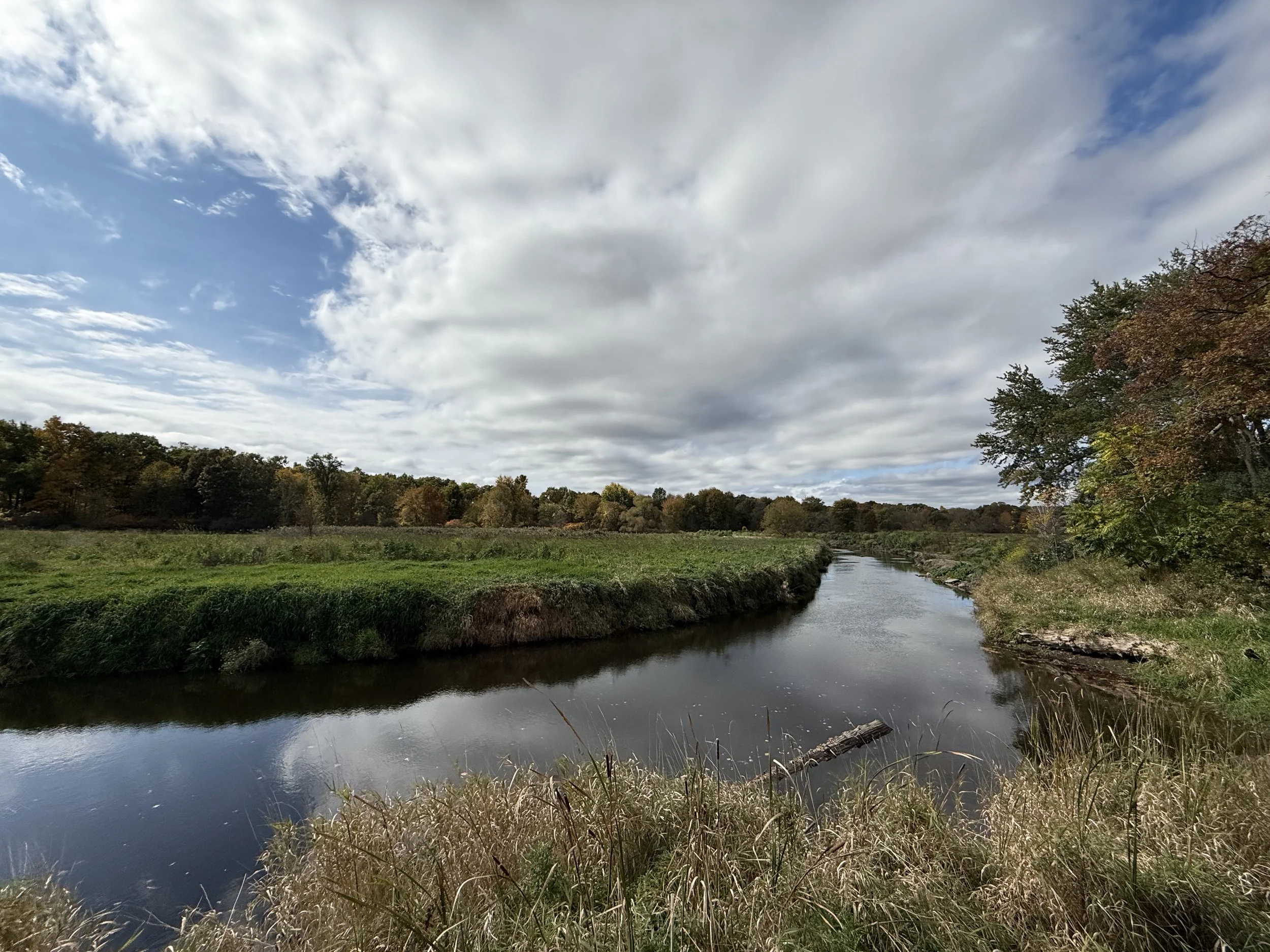 A creek curves around a grassland under a dramatic sky with puffy clouds and blue sky in October.
