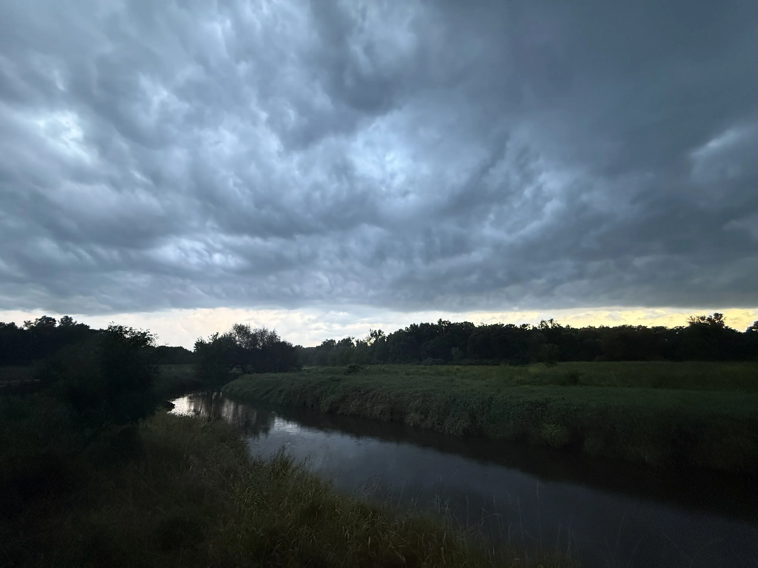 A dramtic dark sky over a creek and grassland edged by woods.