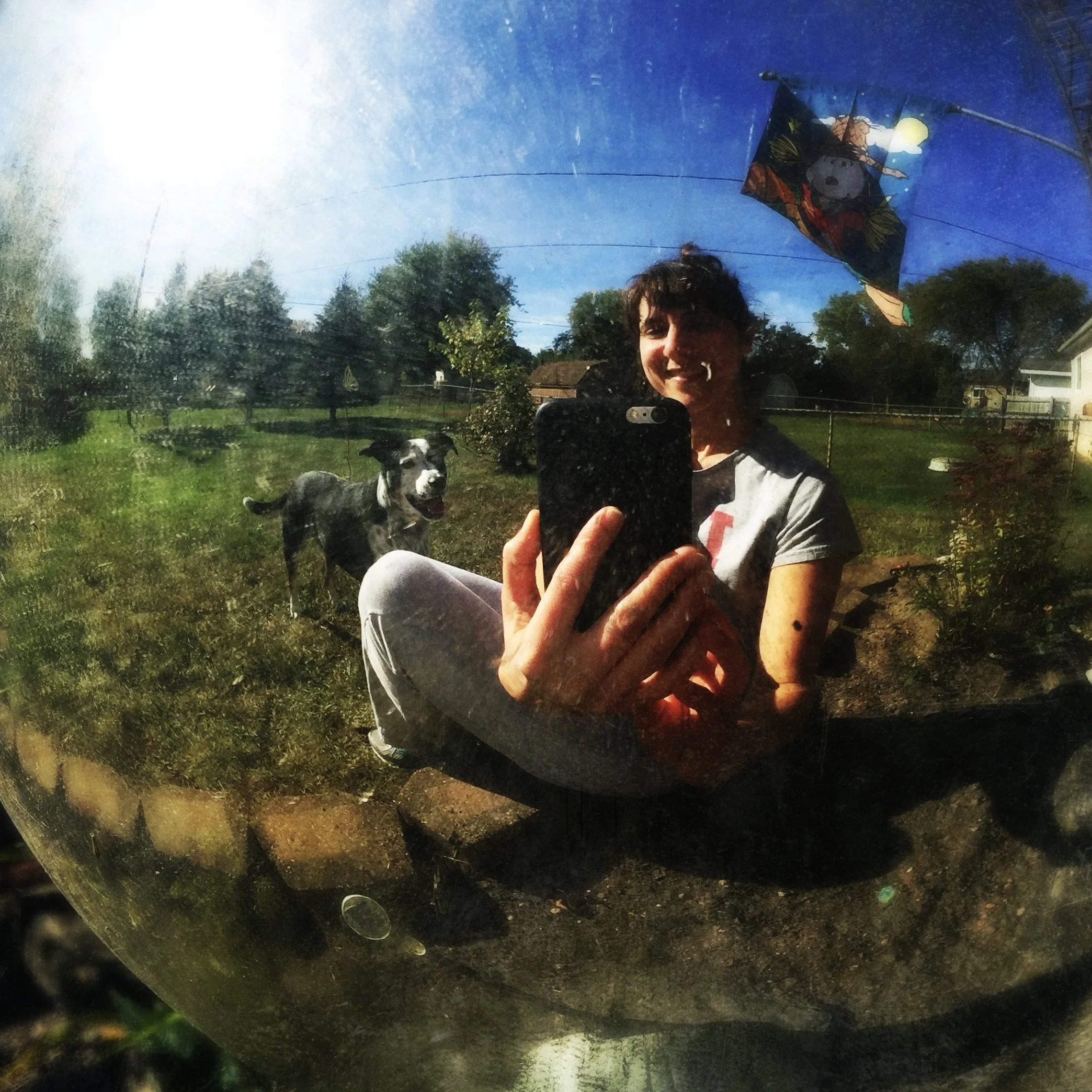 A black and white dog smiles on a sunny day reflected in a garden gazing ball with the person who loves him most.