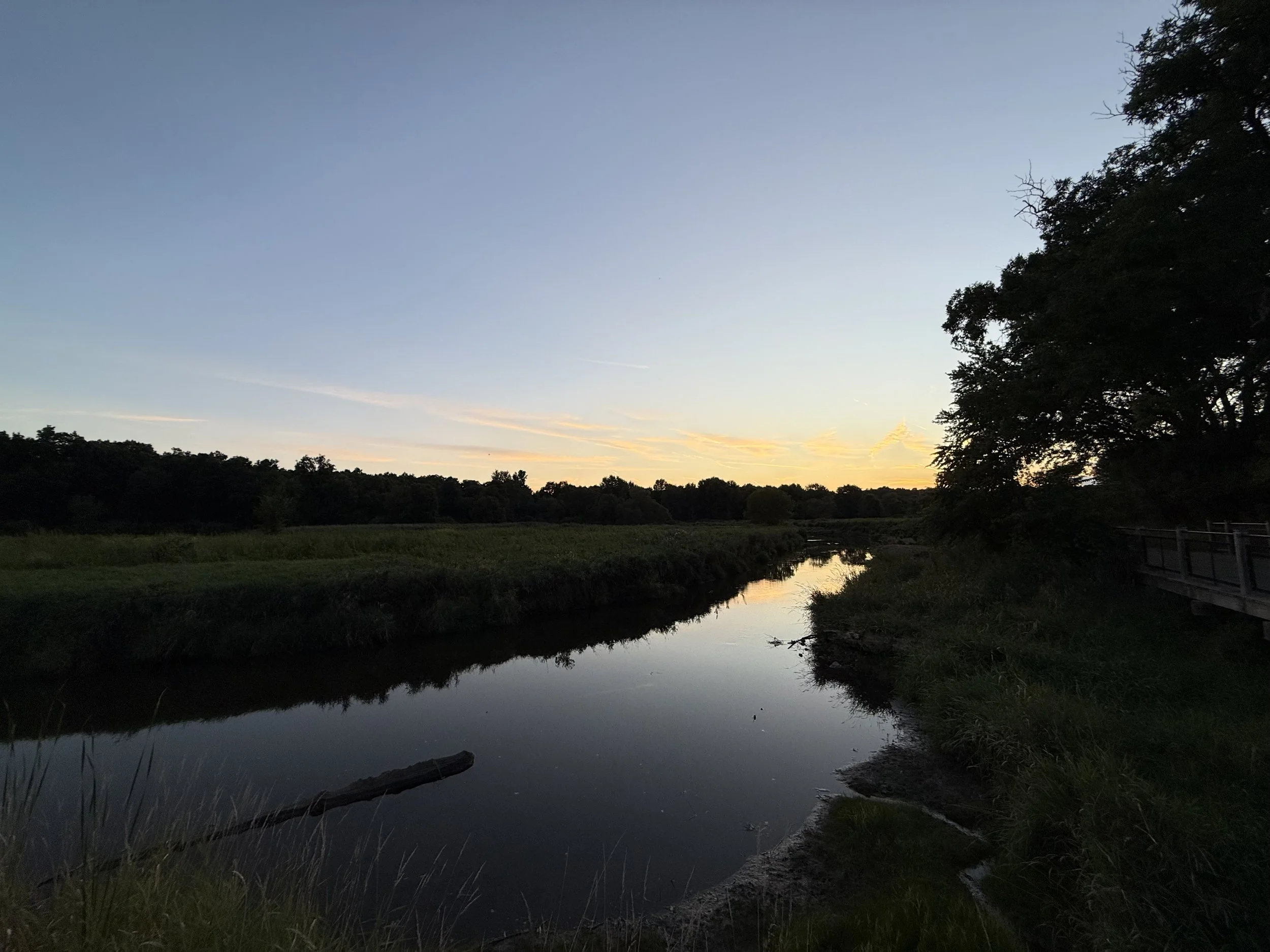 A view of a creek and dramatic clouds over forest and grasslands at nightfall.