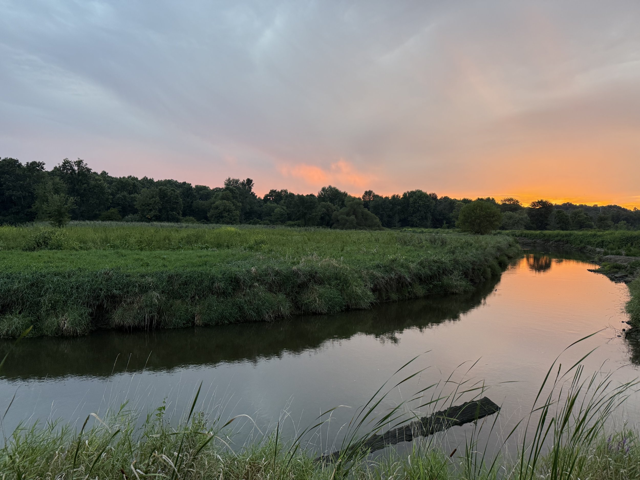 A view of grasslands just after sunset with dramatic orange and pink skies over a winding creek.