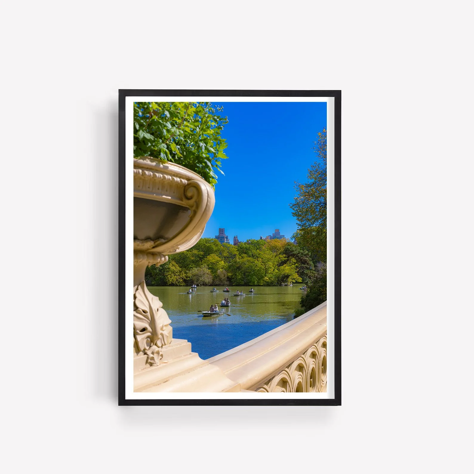Vertical photograph of rowboats on Central Park Lake viewed through the ornate stone balustrade of Bow Bridge, with green trees and the Manhattan skyline beyond.