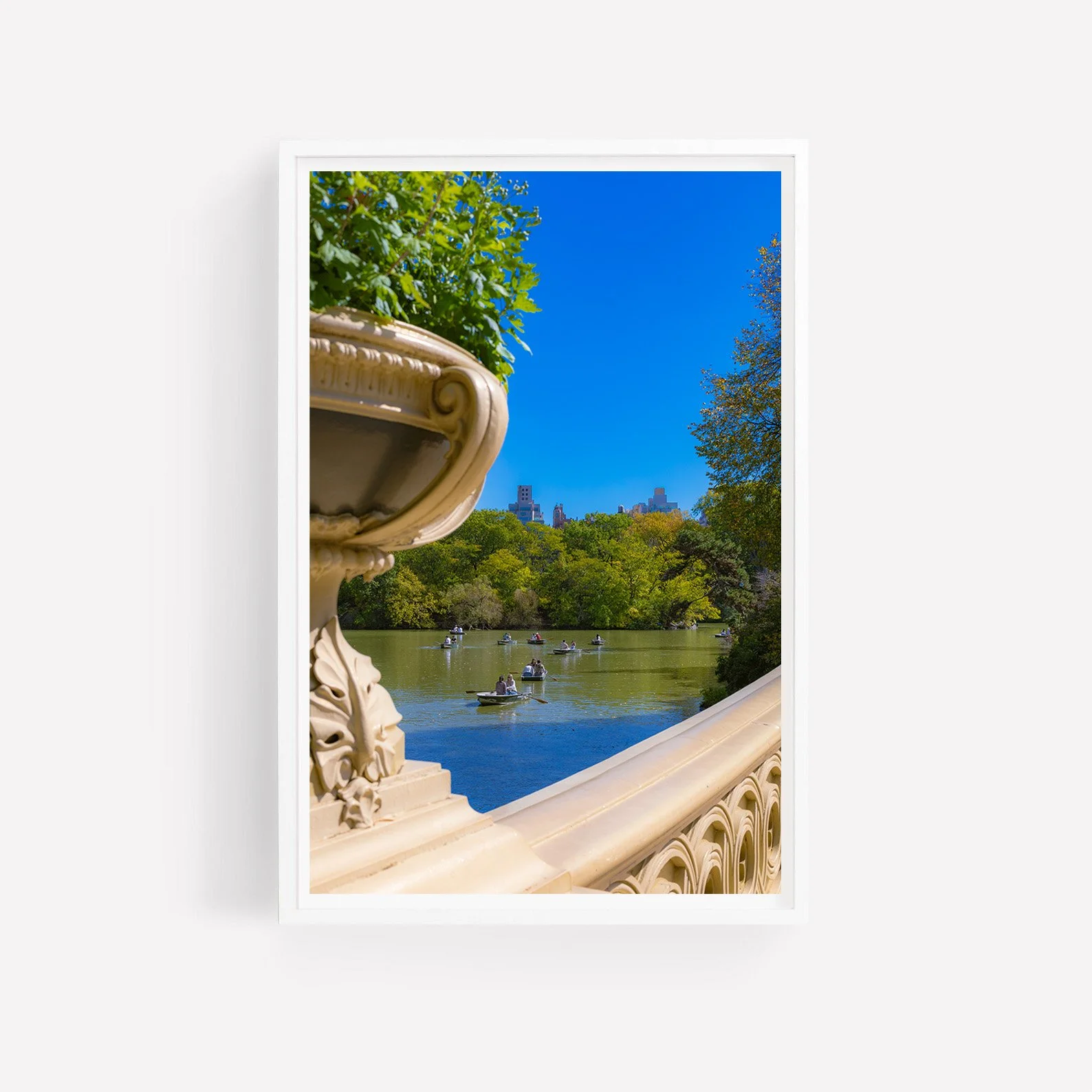 Vertical photograph of rowboats on Central Park Lake viewed through the ornate stone balustrade of Bow Bridge, with green trees and the Manhattan skyline beyond.