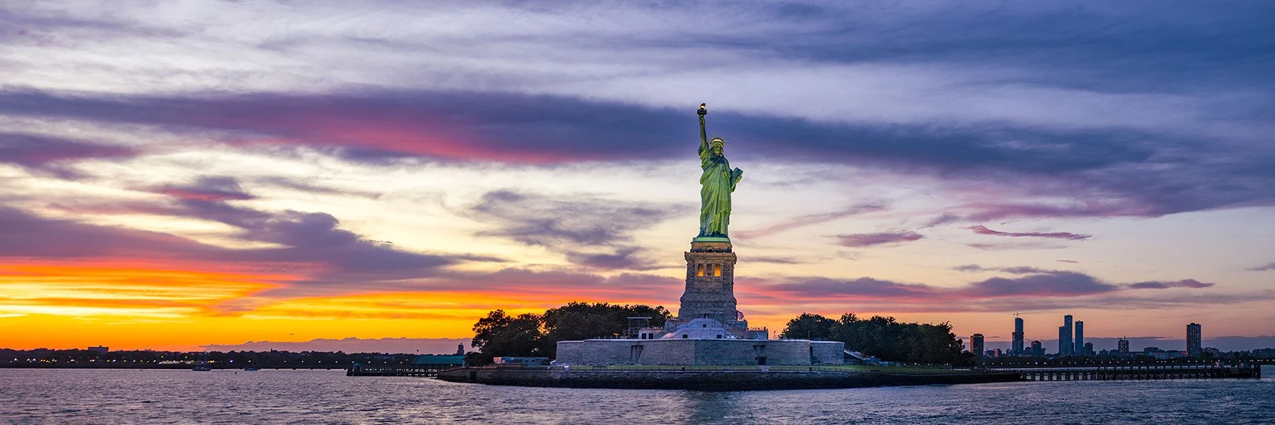 Panoramic fine art photograph of the Statue of Liberty at sunset in New York Harbor, with warm golden and pink tones across the sky and water.