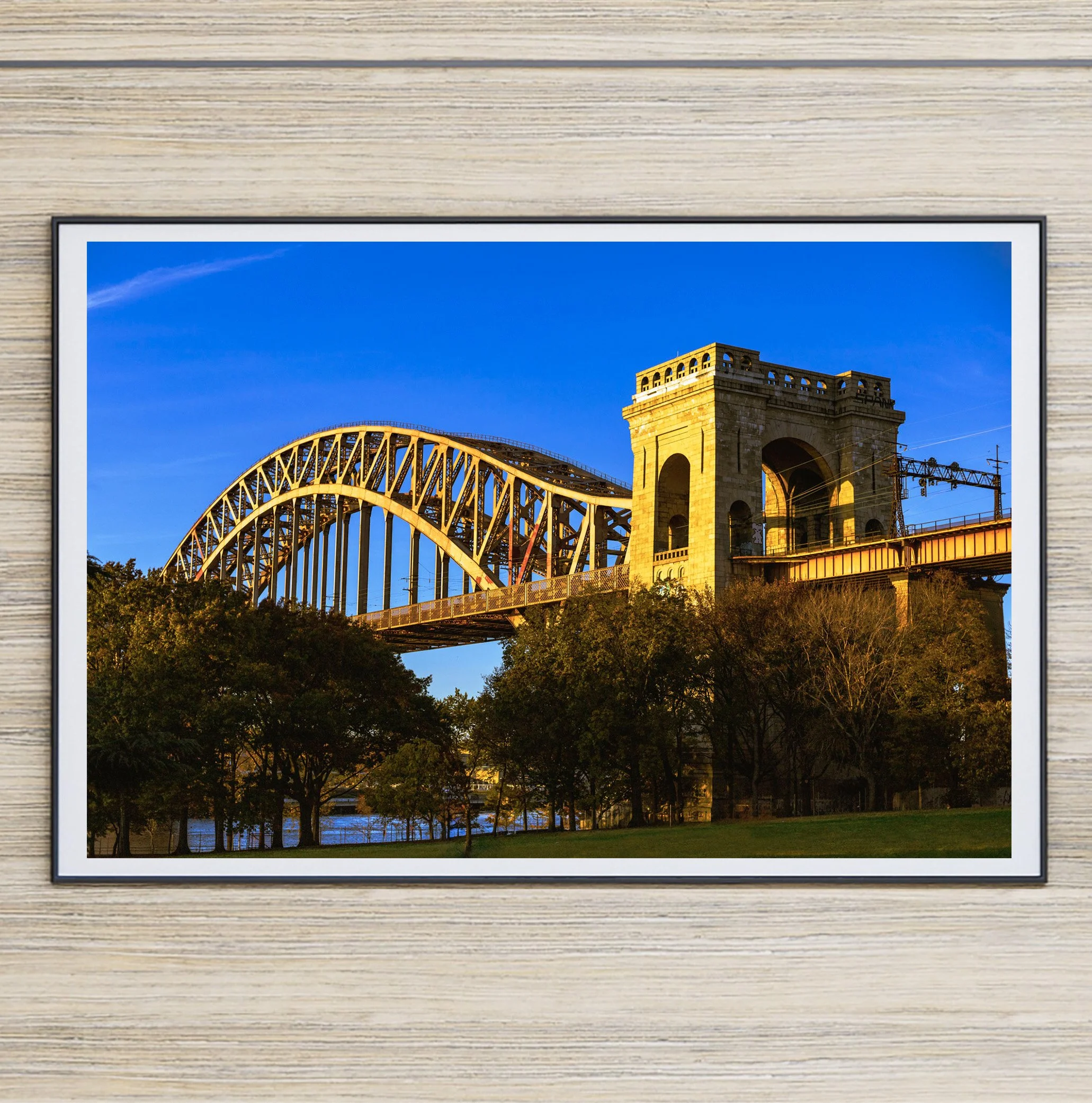 Fine art photograph of the Hell Gate Bridge in Astoria, Queens at sunset, glowing golden above the trees and lawn of Astoria Park beneath a vivid blue evening sky.