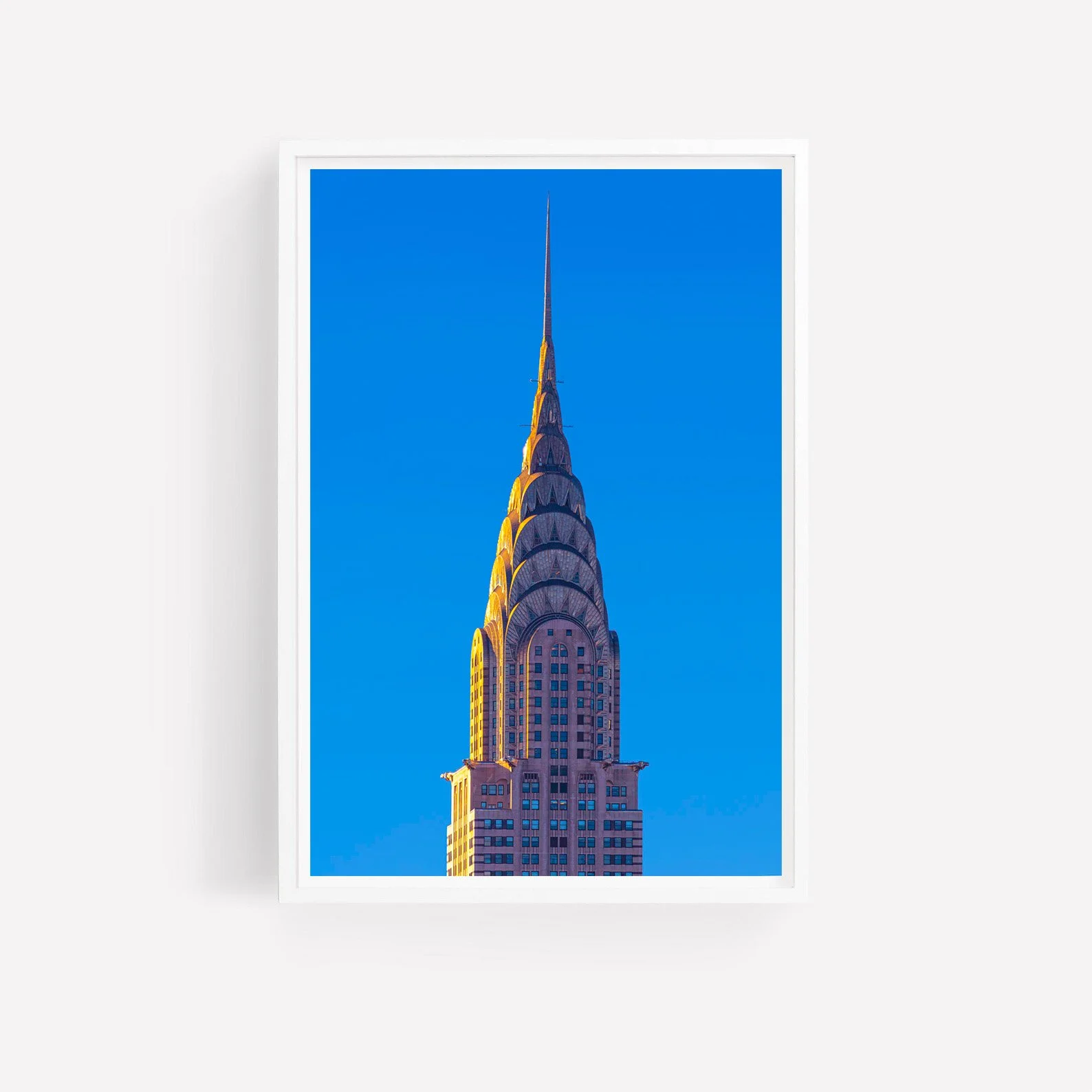 Vertical photograph of the Chrysler Building's iconic Art Deco crown and pointed spire, illuminated by warm sunlight against a vivid blue New York City sky.