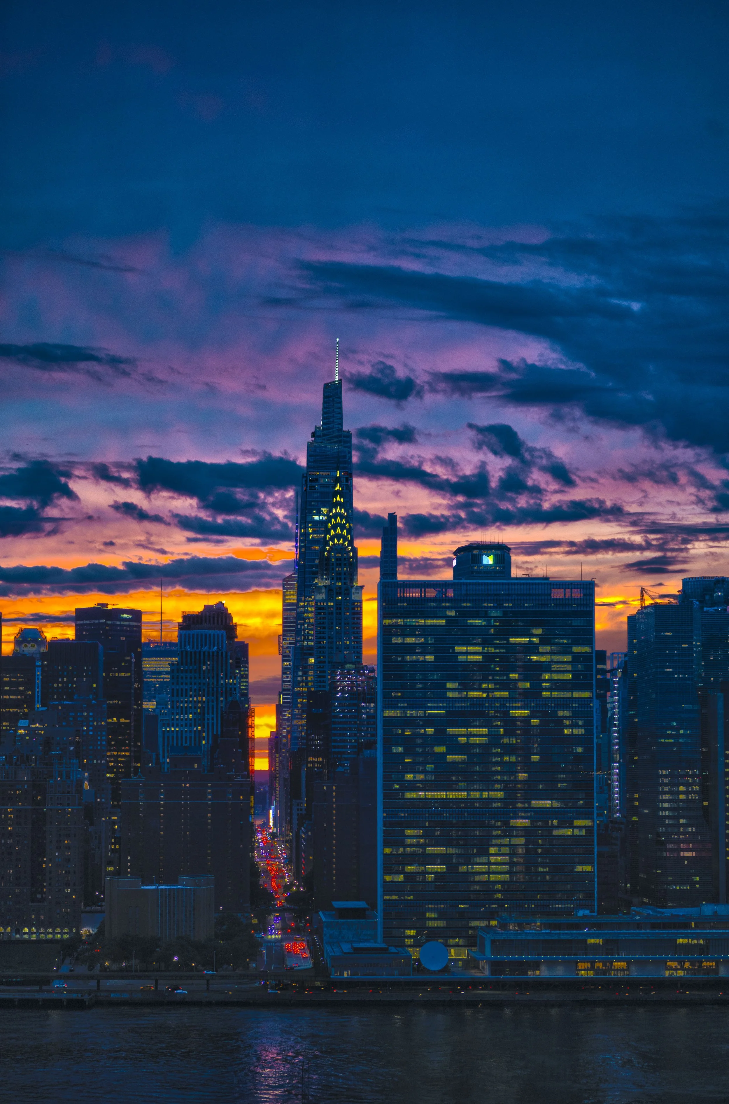 Colorful sunset over Manhattan featuring the illuminated Chrysler Building, the UN Headquarters, the East River, and 42nd Street leading toward Times Square in New York City.