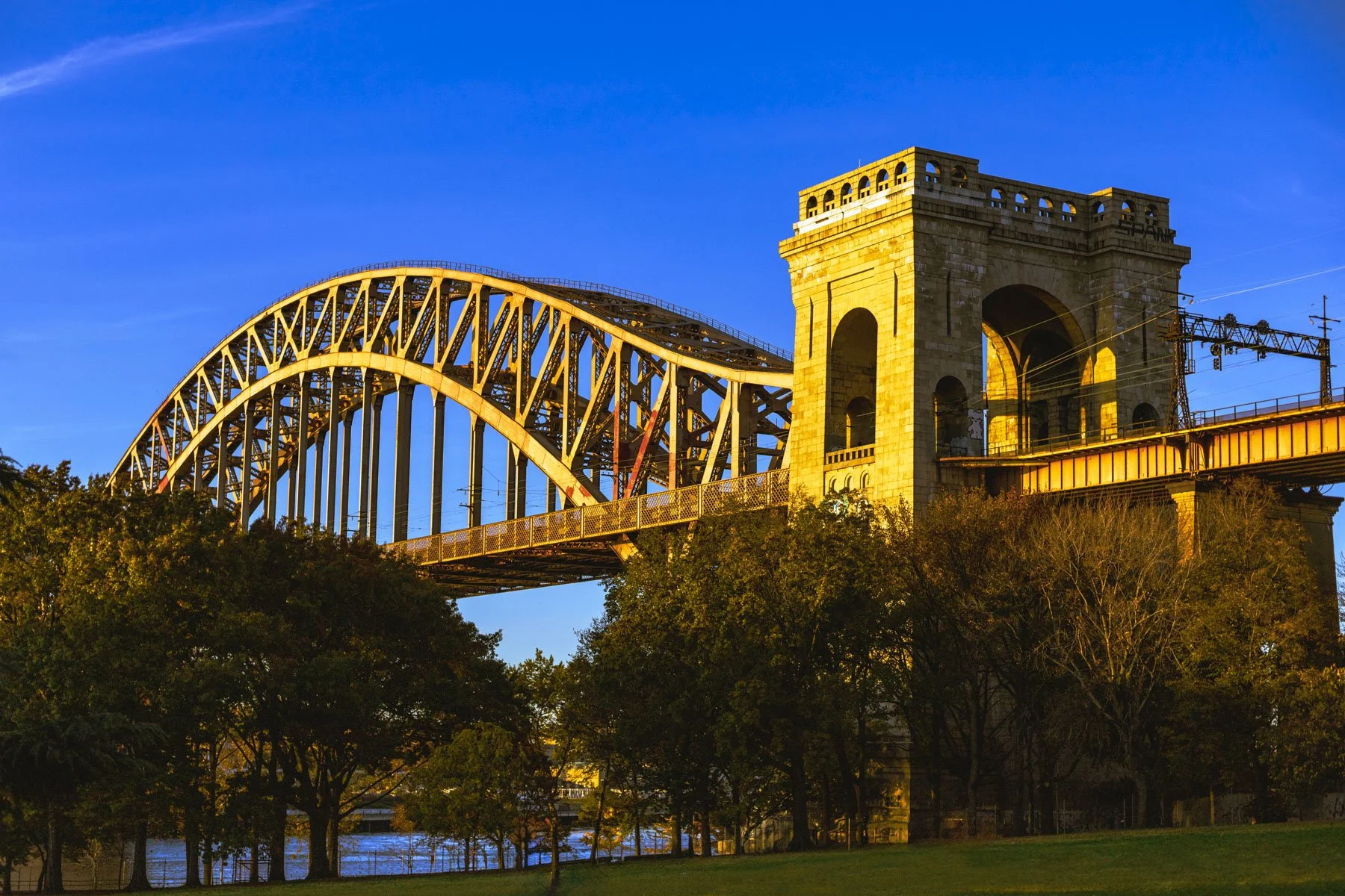 Fine art photograph of the Hell Gate Bridge in Astoria, Queens at sunset, glowing golden above the trees and lawn of Astoria Park beneath a vivid blue evening sky.