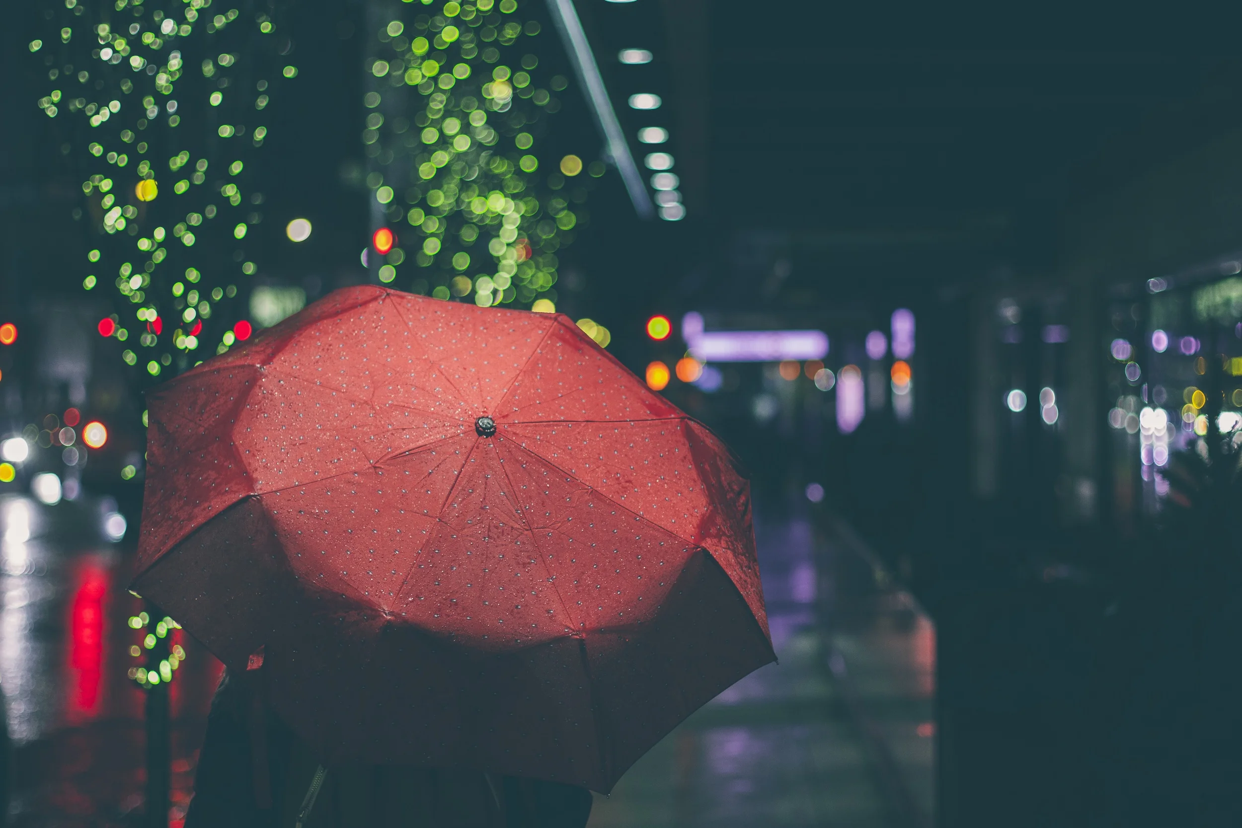 Picture of person with red umbrella walking down a nighttime city street lit with twinkle lights