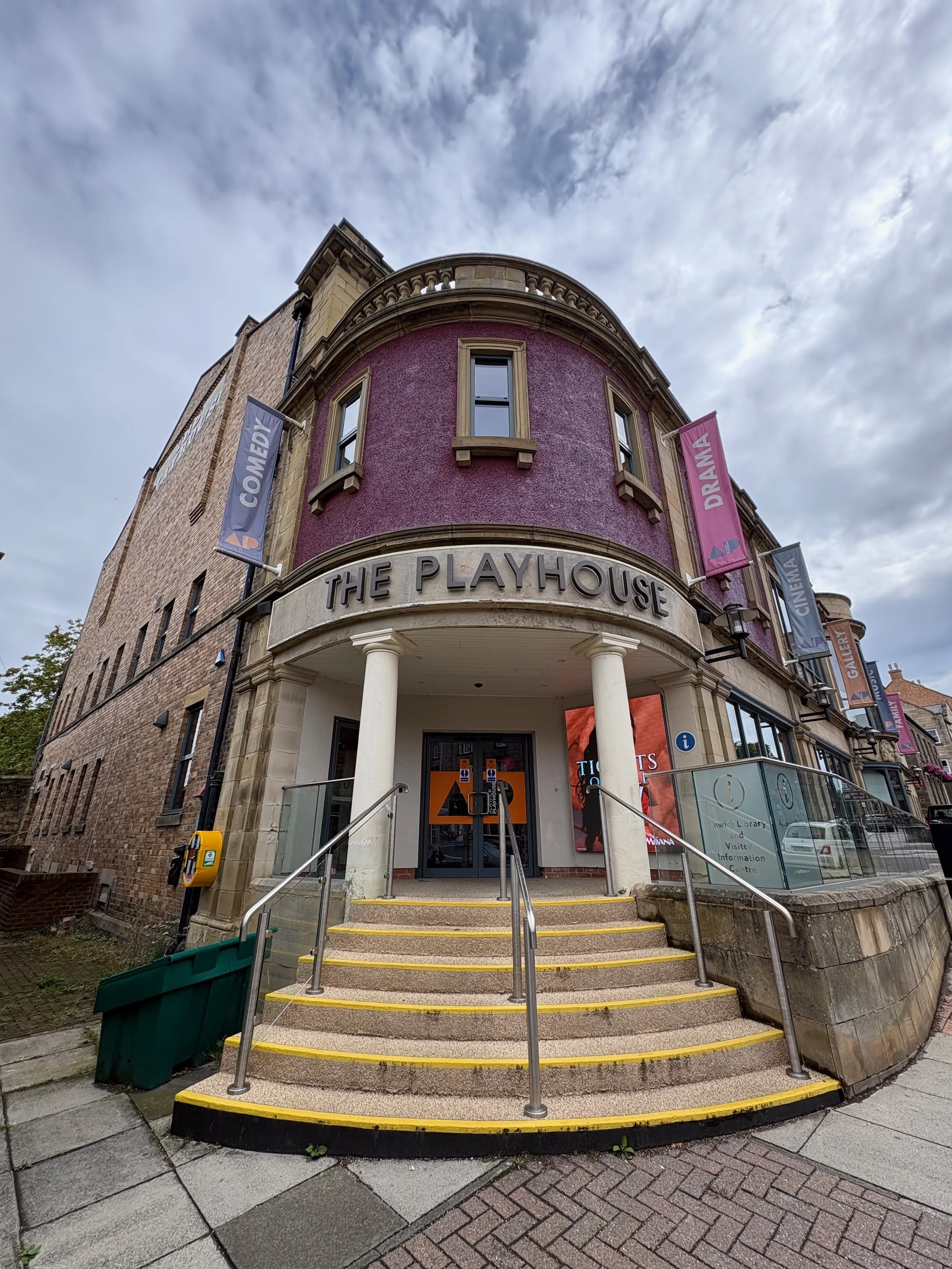 Entrance to The Playhouse, a building with a pink exterior and columns, with banners that read 'Comedy,' 'Drama,' and 'Gallery,' and a step staircase with yellow safety borders.